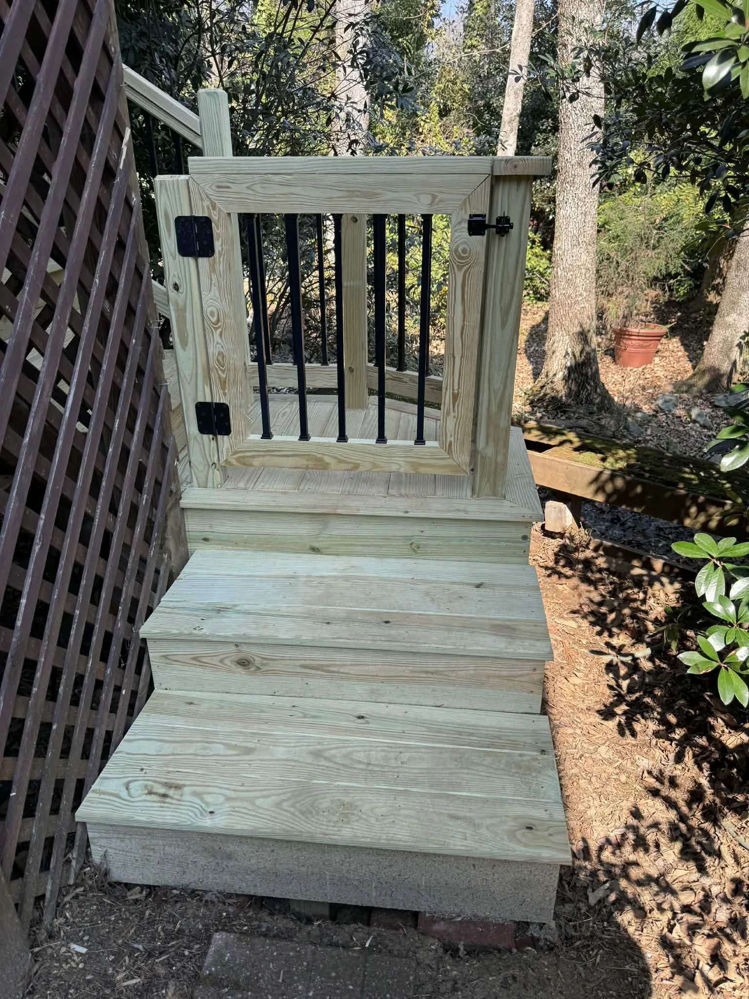 A small wooden deck with a gate, located outdoors in a garden, surrounded by trees and plants.