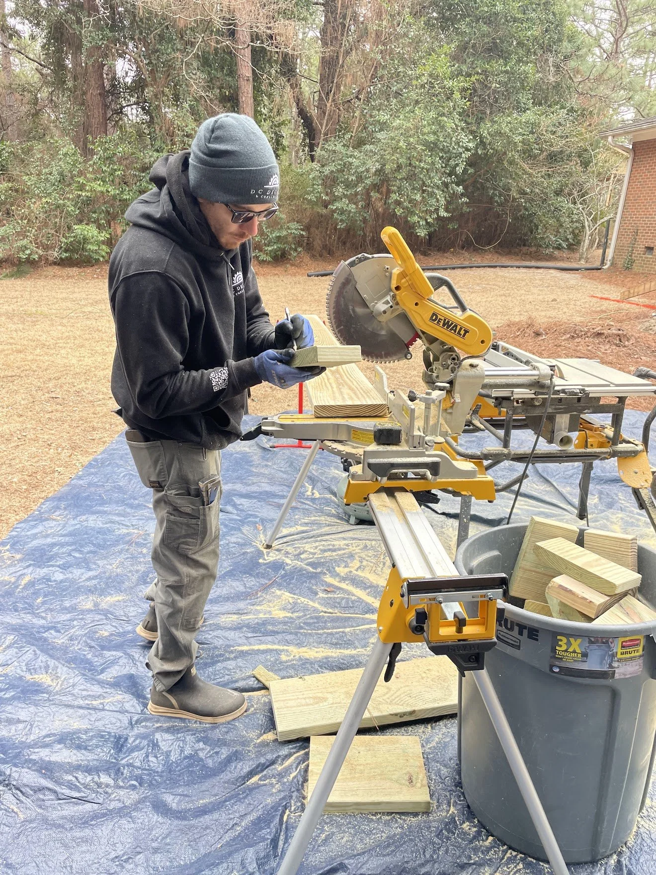 A man wearing a gray beanie and sunglasses marks measurements on a piece of wood with a pencil at an outdoor woodworking project, with a miter saw and other woodworking tools nearby on a work surface and blue tarp on the ground.