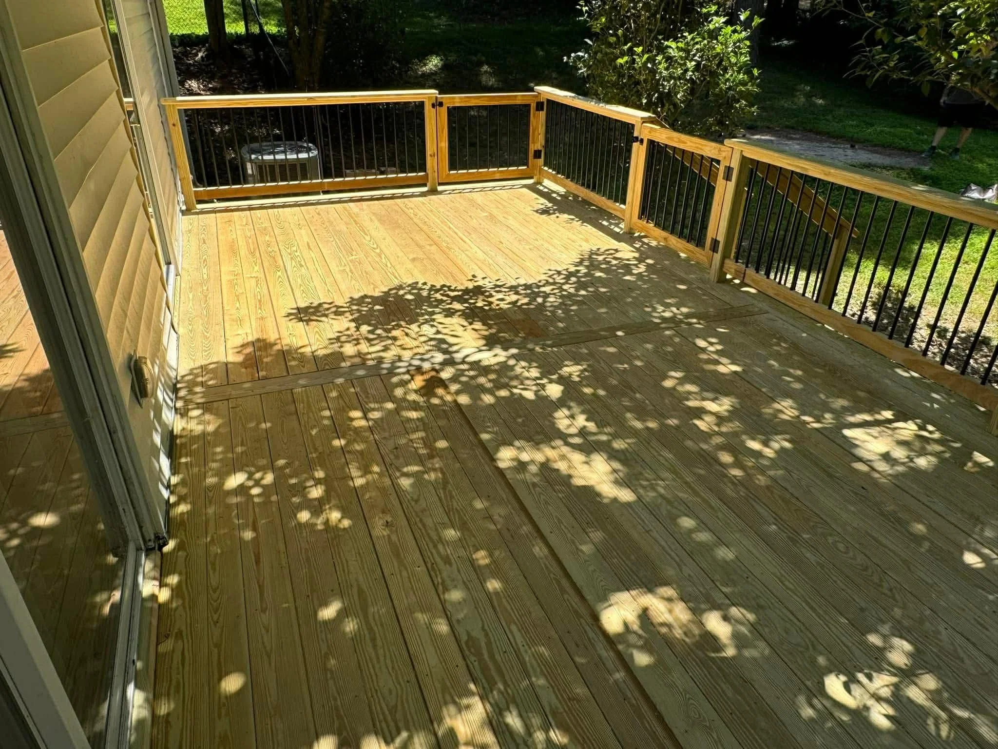 View of a wooden deck with black metal railing, sunlight casting shadows of tree branches on the surface, outdoor greenery visible beyond the deck.