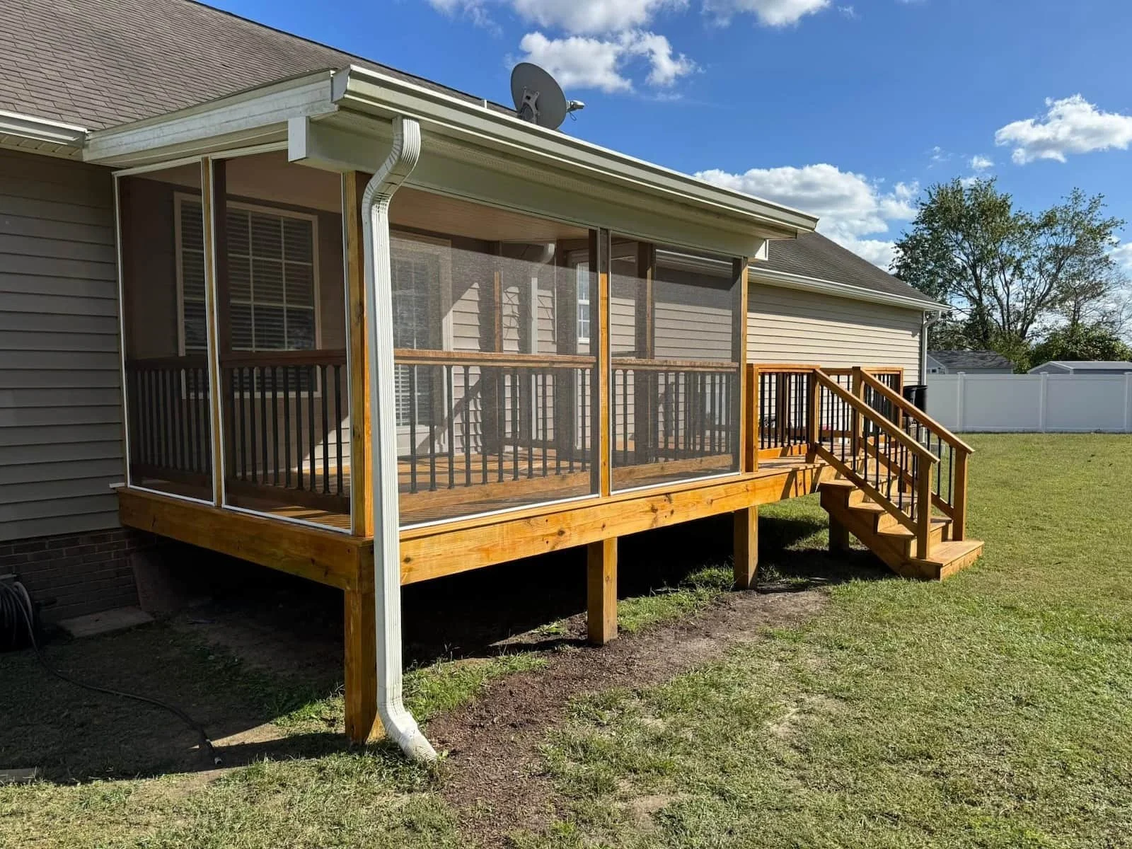 A screened porch attached to a blue house, with black railings, steps, and a door. Inside, there are chairs and a ceiling fan, and outside there's a patch of garden with plants, a small decorative fence, and a propane tank.