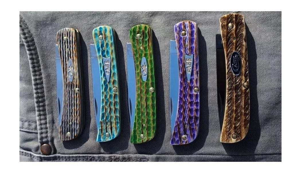 Five colorful pocket knives with textured handles in black, blue, green, purple, and brown, arranged on a gray fabric surface.
