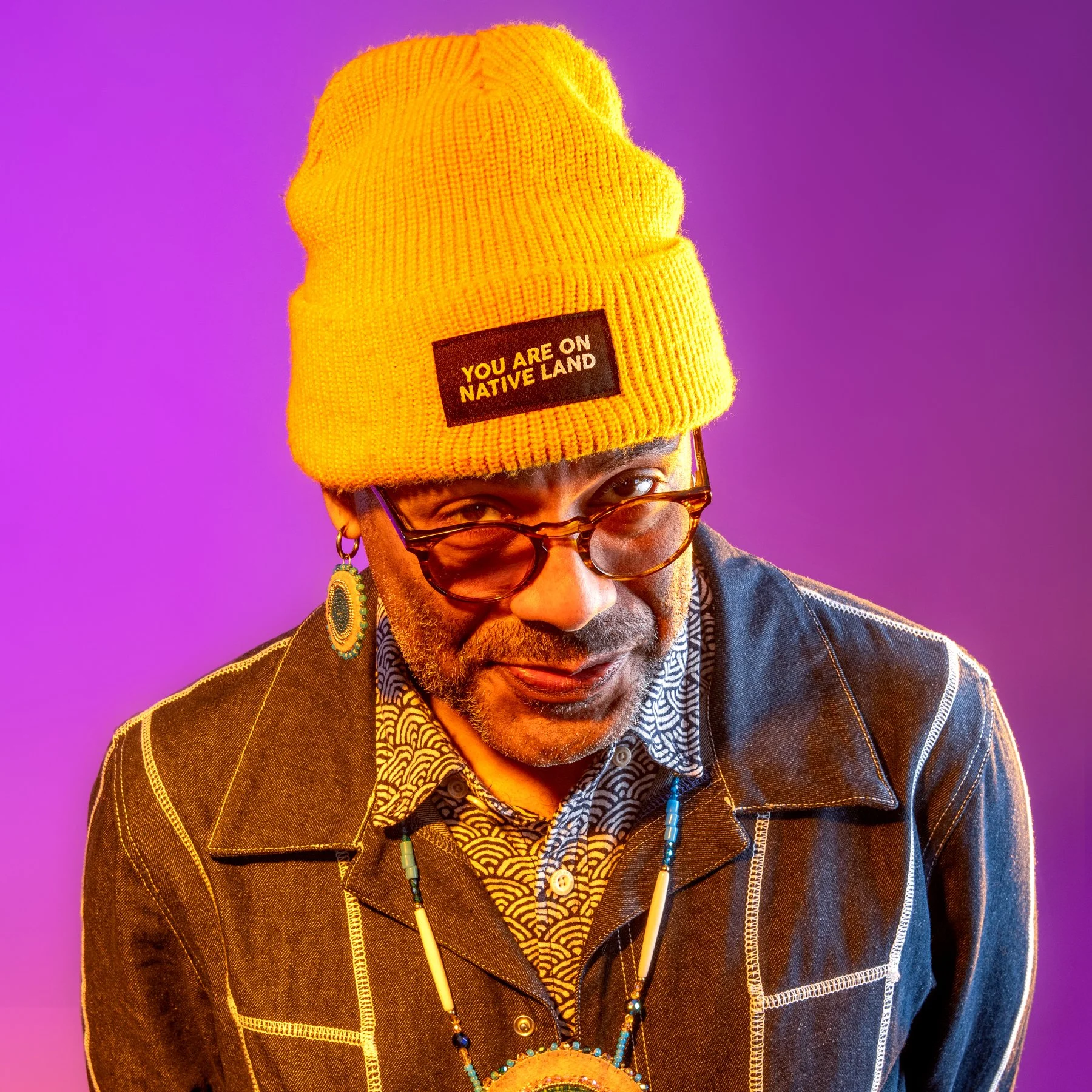 Profile photo of Carlos Kareem Windham against a purple backdrop. They are wearing a yellow knit with a tag that reads "you are on native land" and a brown jacket with white seams.
