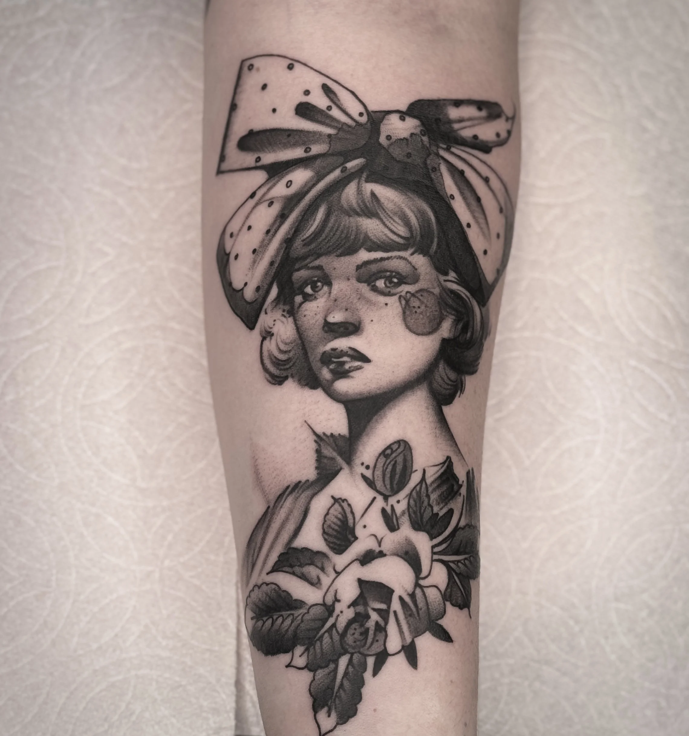 Stylized portrait tattoo of a woman with a large polka-dot bow and soft expression. Freckled, floral, and timeless.