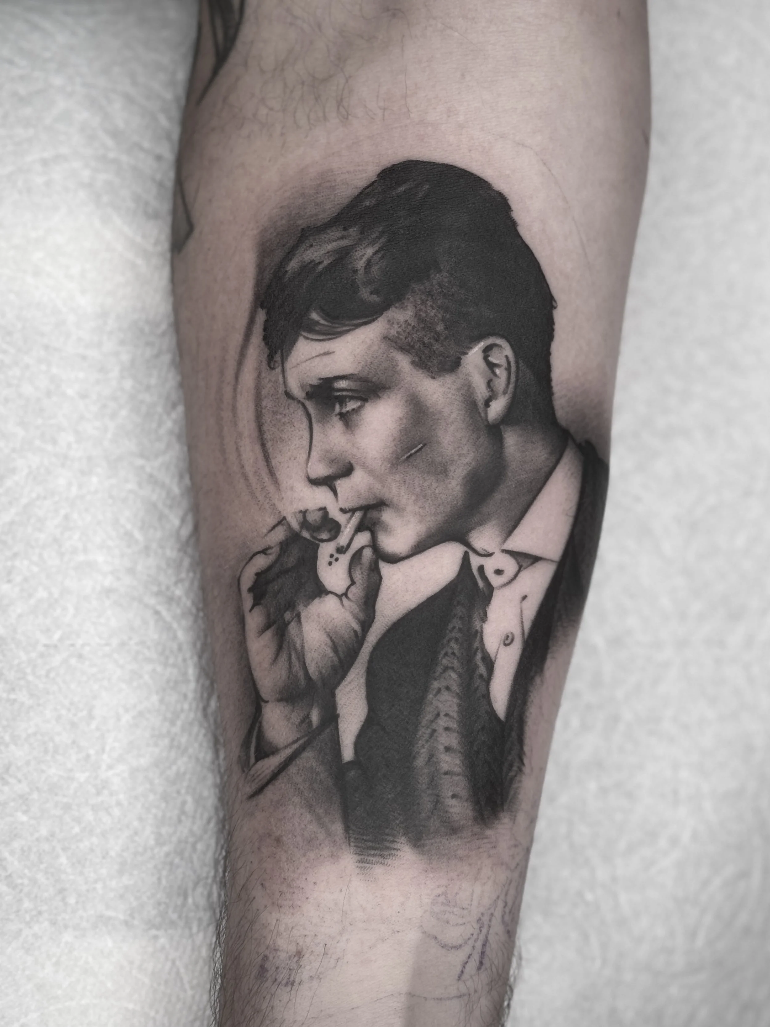 Black and grey portrait tattoo of Thomas Shelby (Cillian Murphy) from Peaky Blinders, in profile, holding a cigarette. Soft cinematic shading with stylized smoke effect.