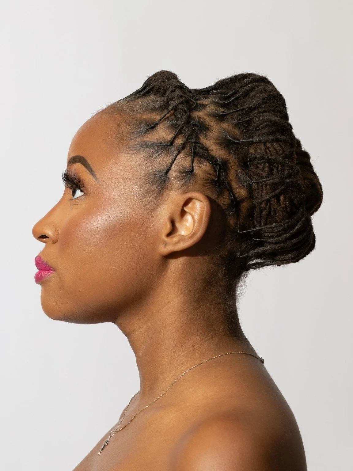 Profile view of a woman with intricate braided hairstyle, featuring dreadlocks pulled back into a bun, wearing a necklace and lipstick.