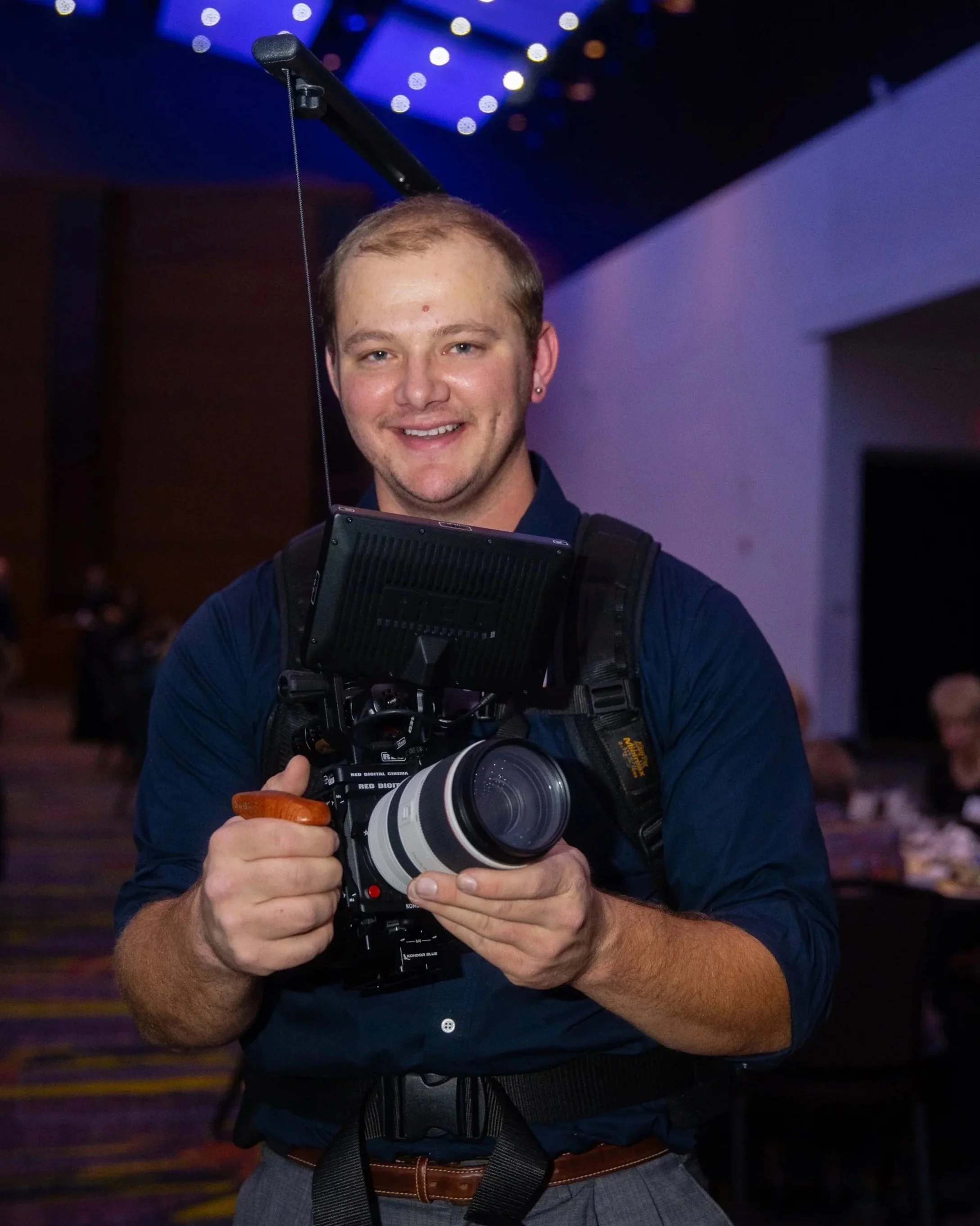A man with long hair and beard, wearing headphones, is looking at his phone in a dimly lit, industrial-style room with exposed wires and a neon sign in the background.
