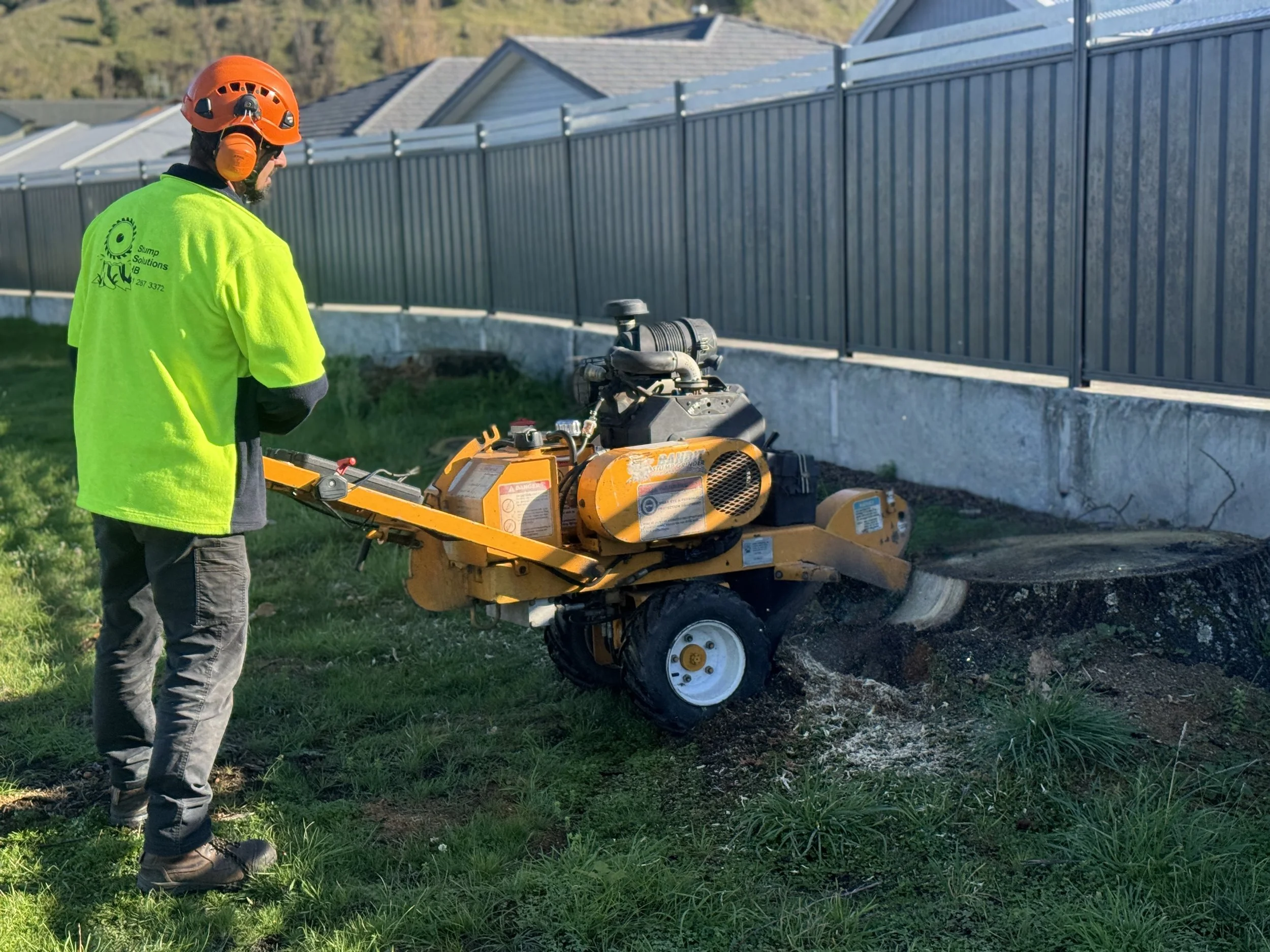 Franek (owner) in a neon yellow safety shirt and orange helmet operating a yellow stump grinder on a tree stump in a grassy yard next to a metal fence.
