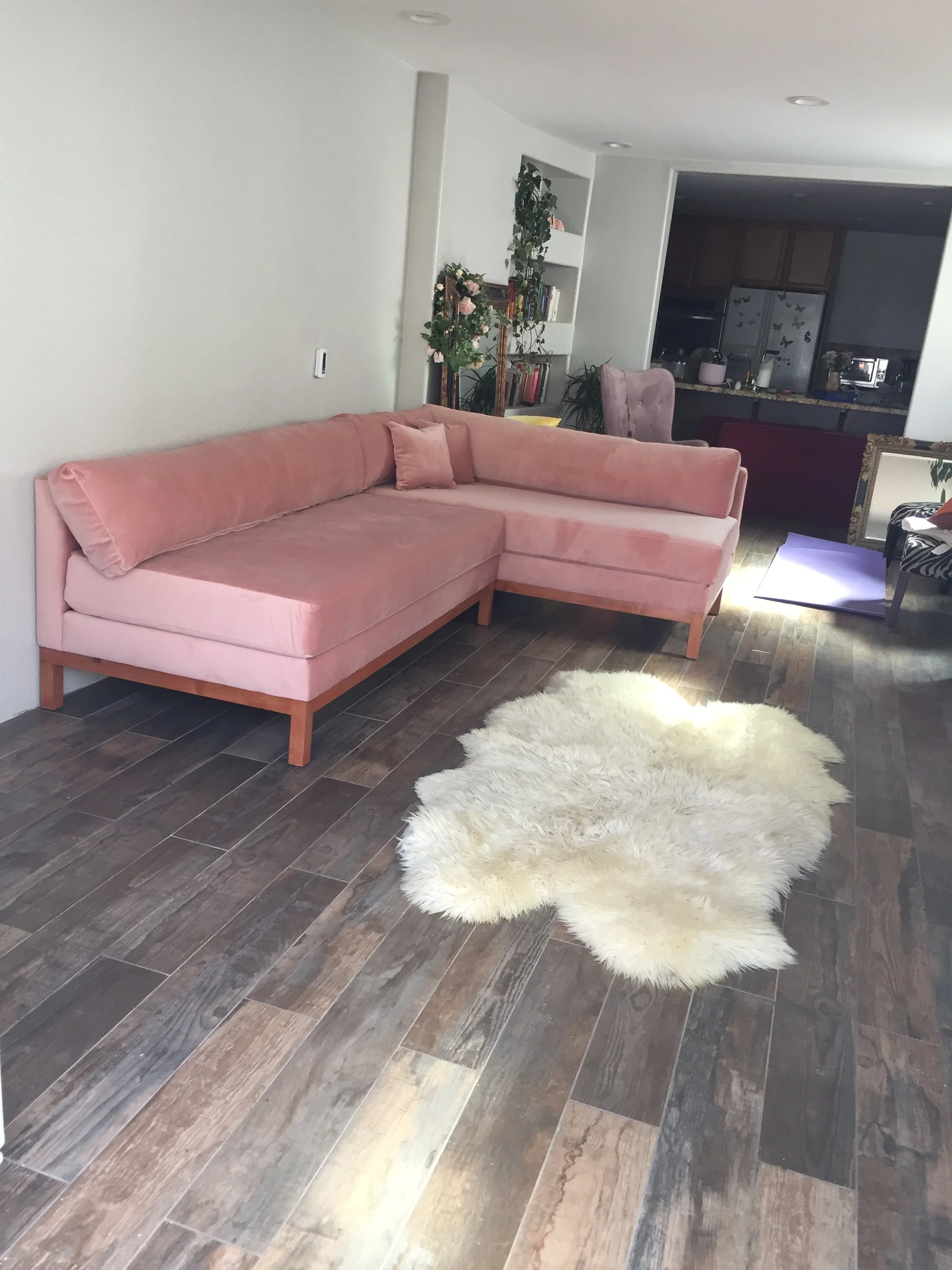 Living room with a pink sectional sofa, white shag rug, and wooden flooring. Background includes a bookshelf, plants, a purple chair, and a view into the kitchen area.