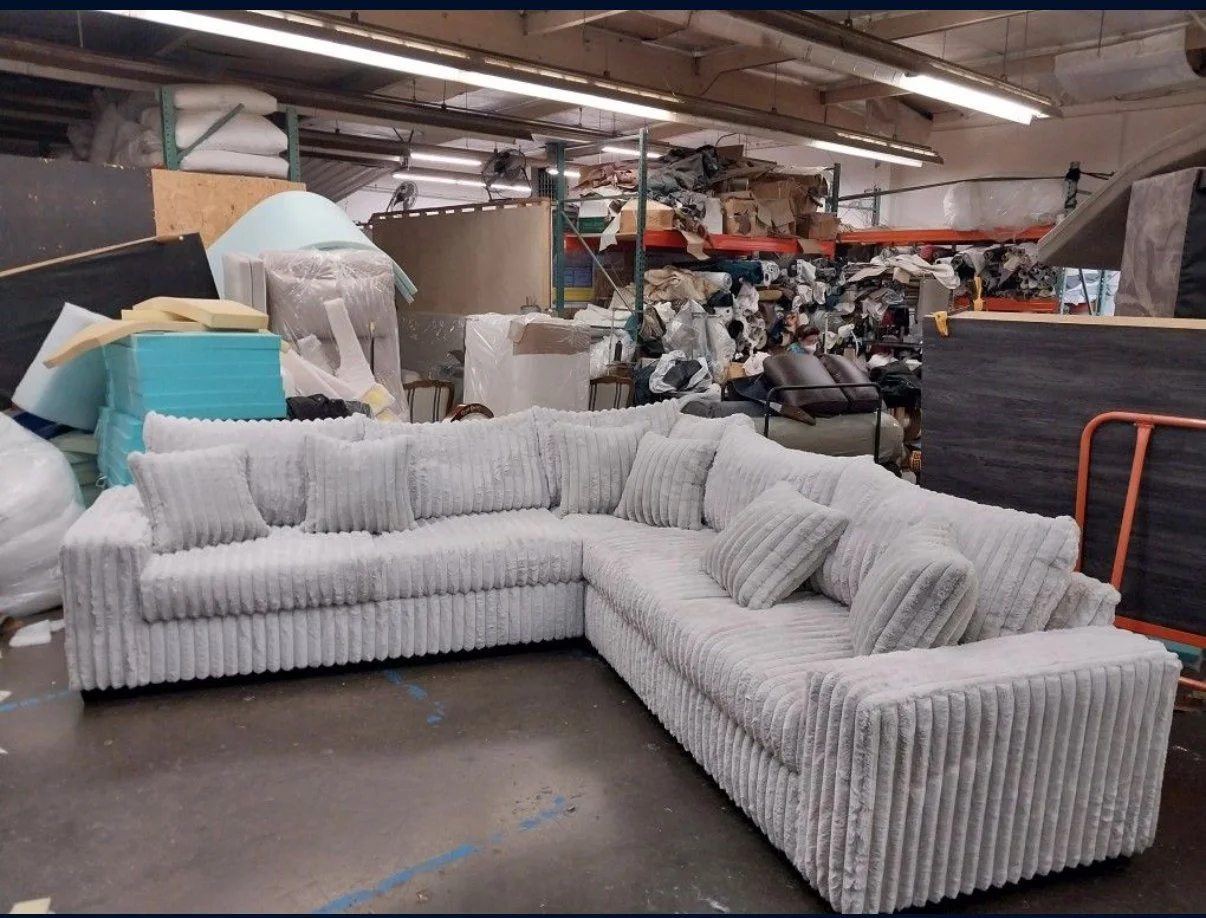 A light-colored, striped L-shaped sectional sofa with matching pillows, placed in a warehouse or storage area filled with various furniture and fabric items.