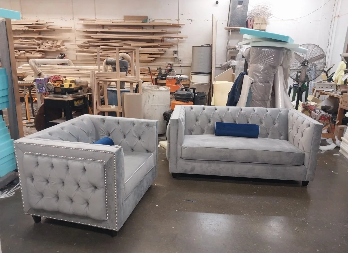 Two light gray tufted sofas with dark wooden legs and blue pillows in a woodworking or upholstery workshop, surrounded by tools, wood pieces, and workshop materials.