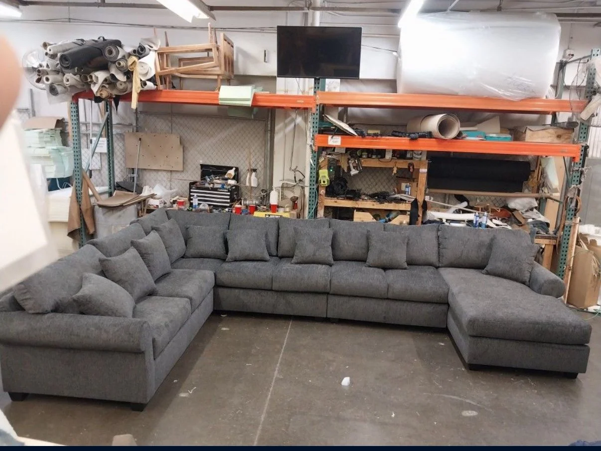 A large gray sectional sofa with multiple cushions is placed in a warehouse or workshop surrounded by various tools, materials, and storage shelves.