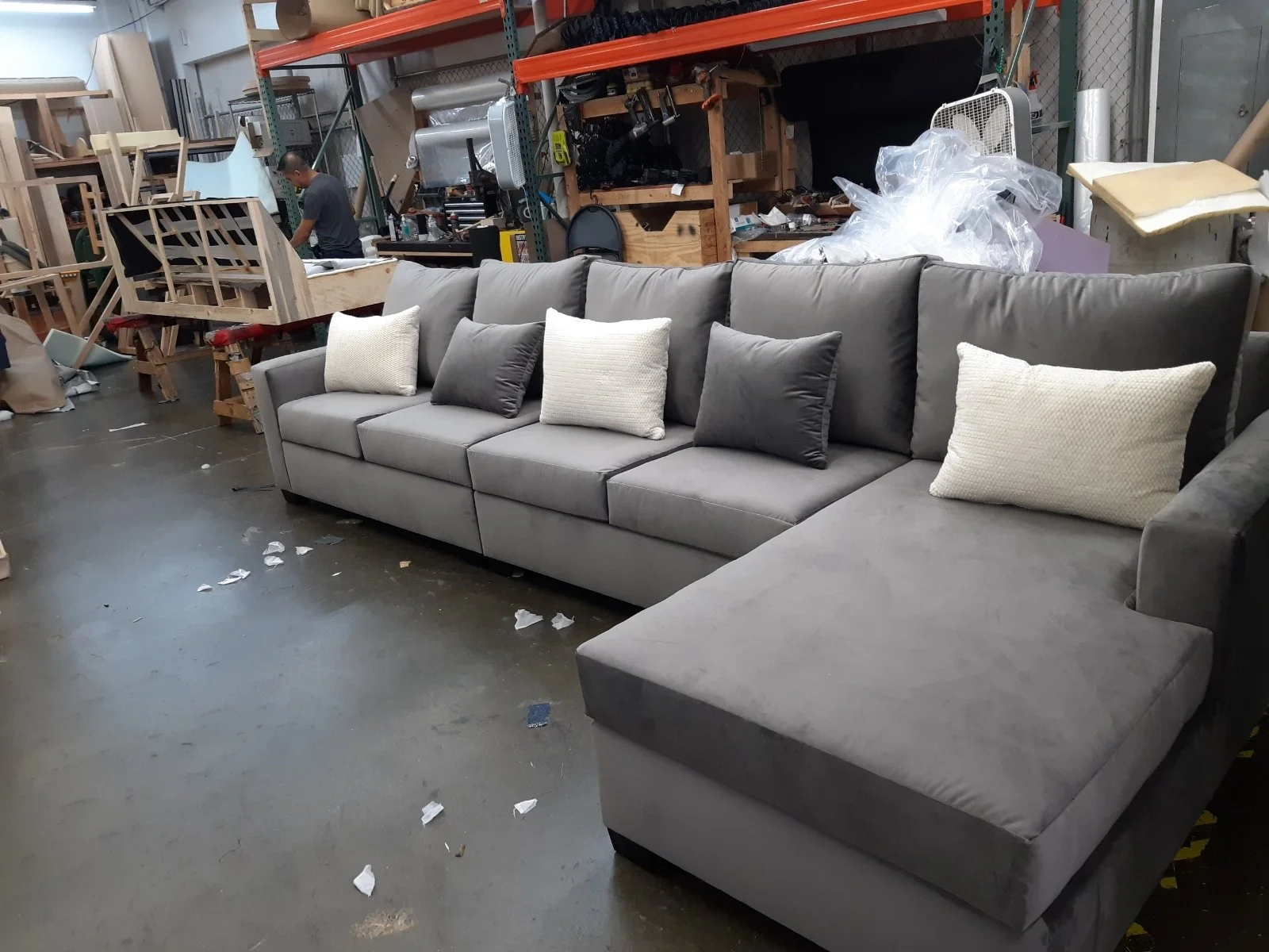 A grey sectional sofa with six throw pillows, four white and two black, in an industrial workspace with wooden furniture and tools in the background.