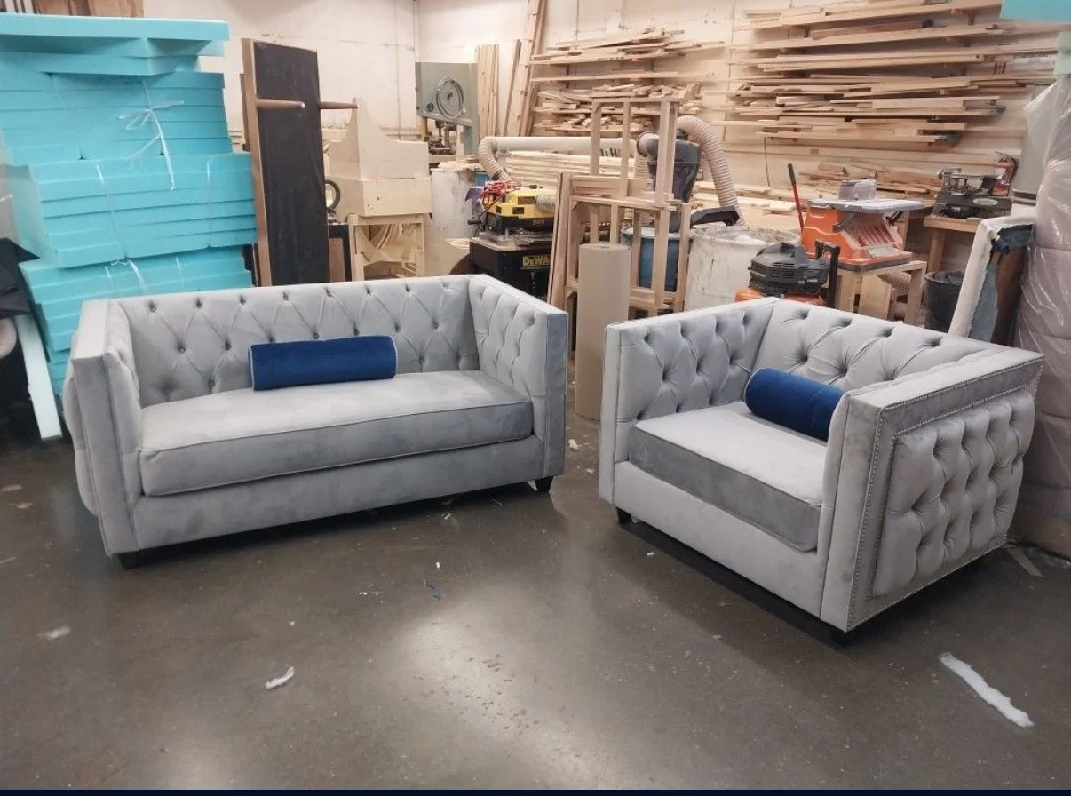Two gray tufted sofas with rolled arms and dark wooden legs, each with a blue cylindrical cushion, in a woodworking workshop with stacked wood and tools in the background.