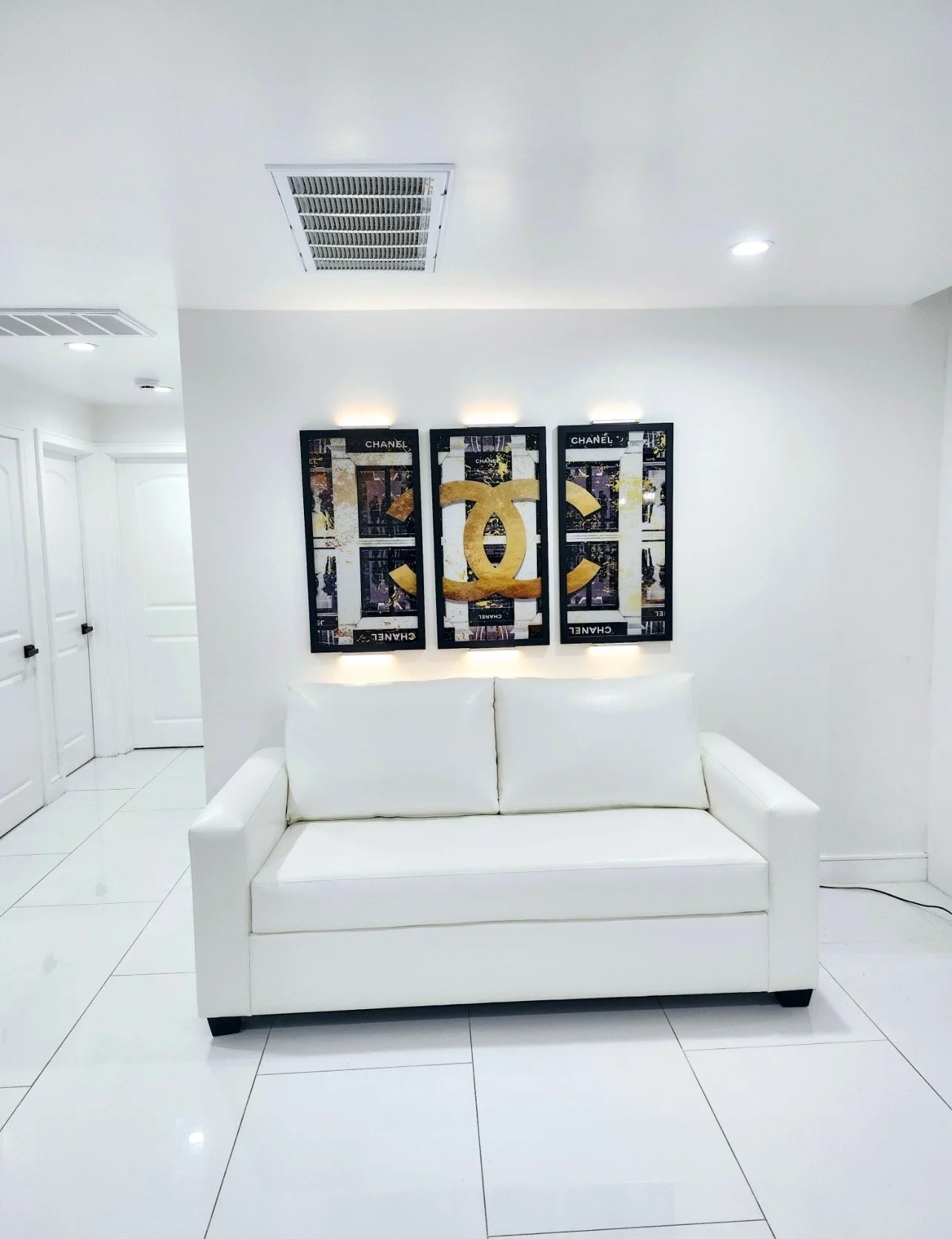 Modern living room with white walls and floor, featuring a white sofa and three framed Chanel artwork pieces with gold and black detailing on the wall above the sofa.