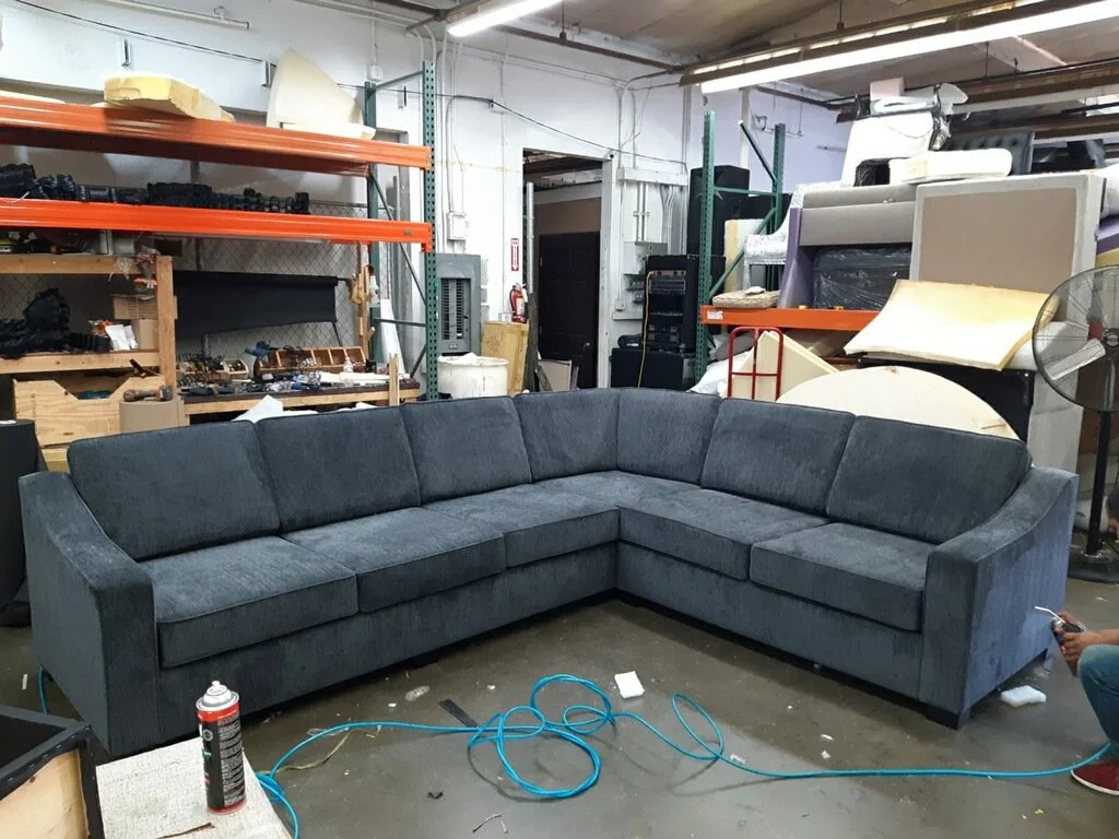 A large L-shaped gray sofa in a warehouse or storage room with shelves holding various items, including foam, cardboard, and tools.