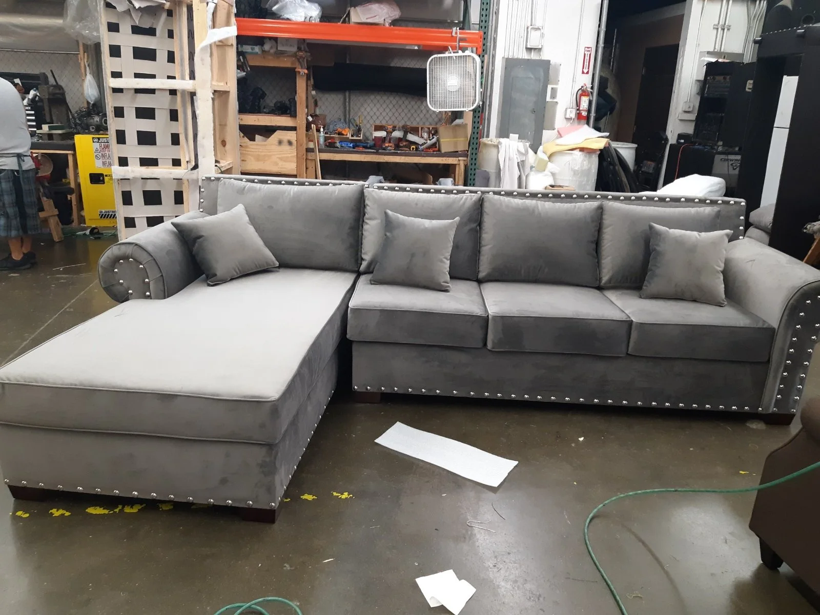 Gray velvet sectional sofa with studded nailhead trim and four matching pillows in a warehouse or upholstery workshop.