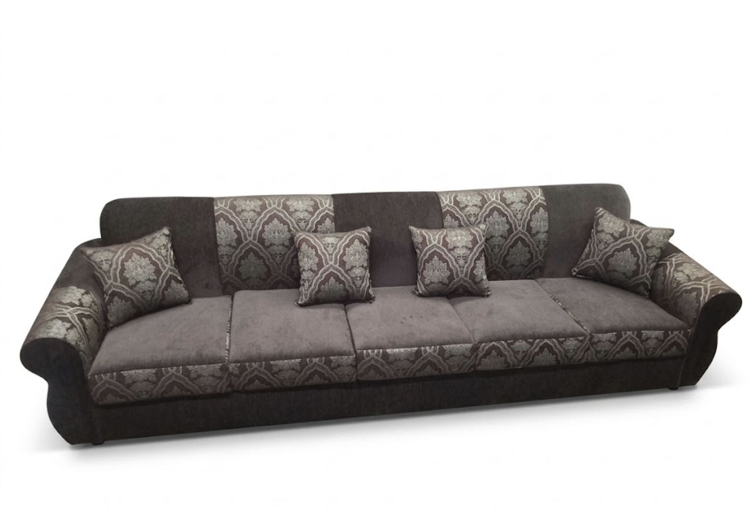 A long, plush sofa with a patterned fabric backrest and armrests, decorated with five matching throw pillows, set against a plain white background.