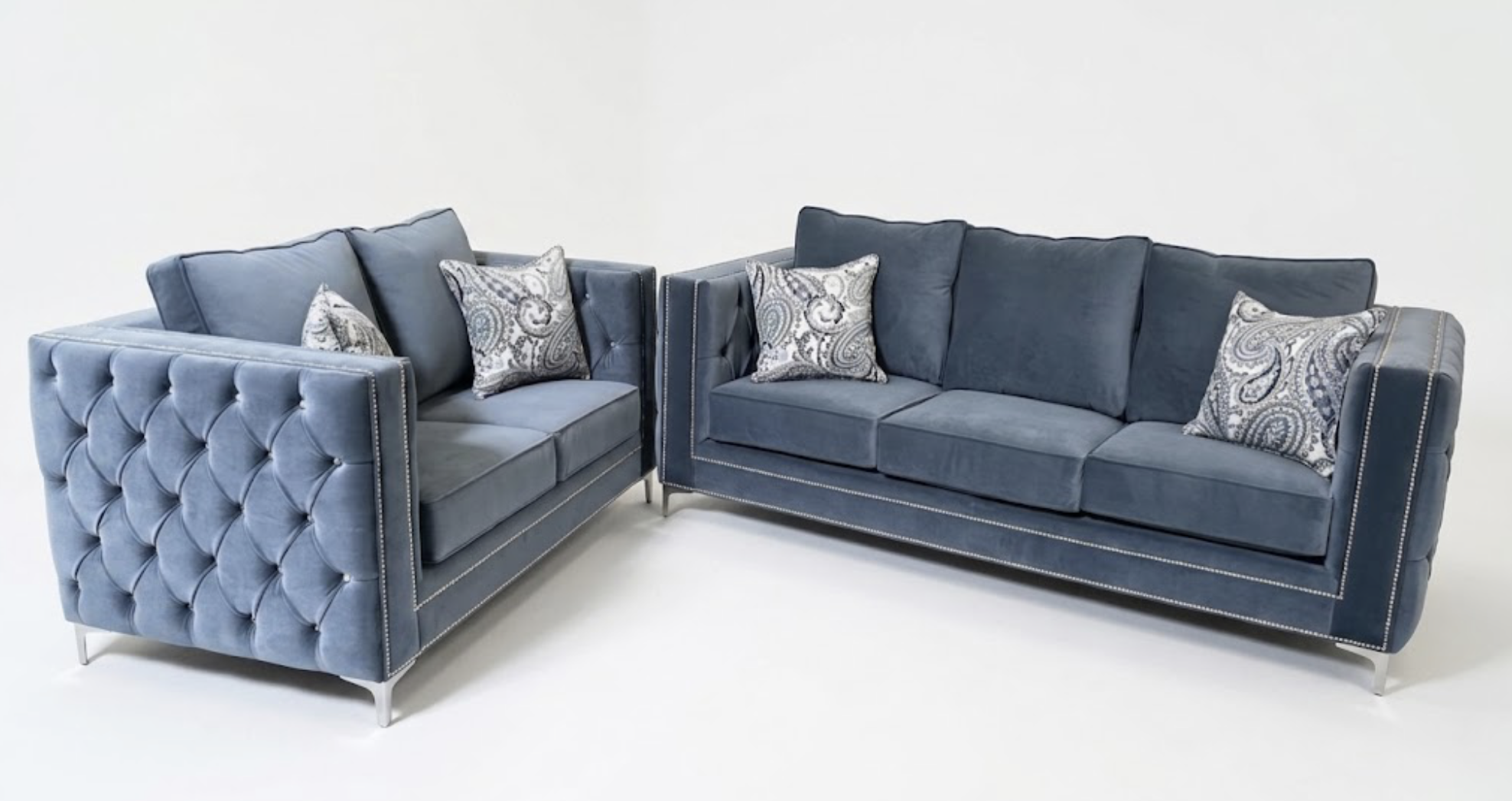Two navy blue velvet sofas, one larger and one smaller, against a plain white background. The sofas have button-tufted sides, nailhead trim, and are decorated with assorted patterned throw pillows.