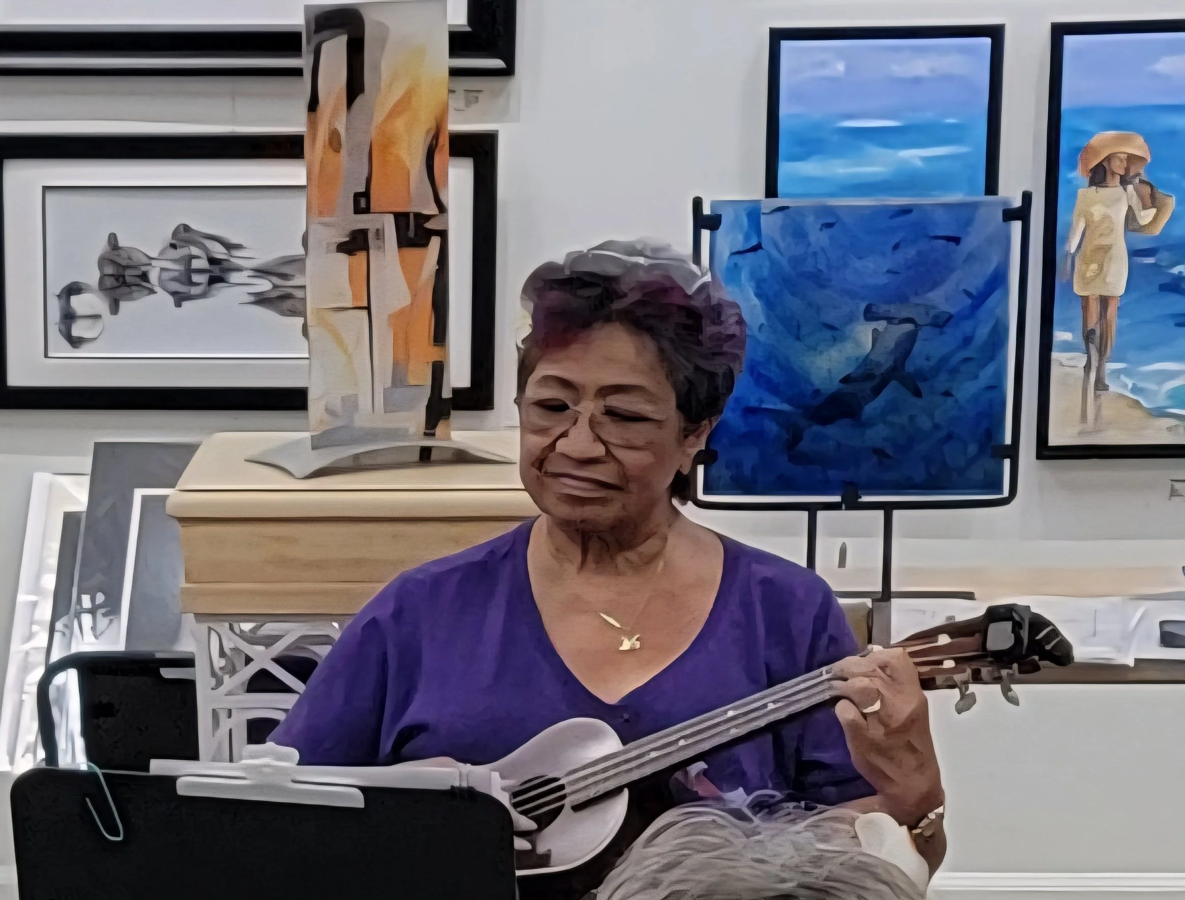 An elderly woman with short gray hair and glasses, wearing a purple top, is playing an acoustic guitar. She is smiling and sitting in front of several framed paintings of sea scenes and a woman in a yellow dress with a sunhat. The environment appears