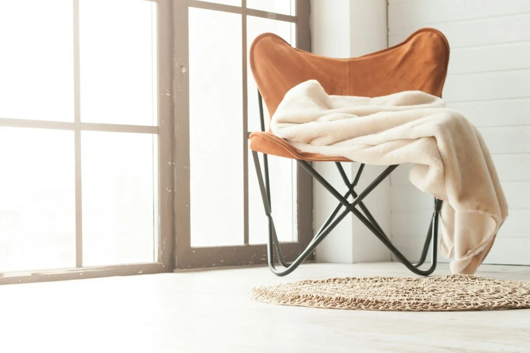 A brown butterfly chair with a cream blanket draped over it, placed on a round woven rug near large windows in a bright room.