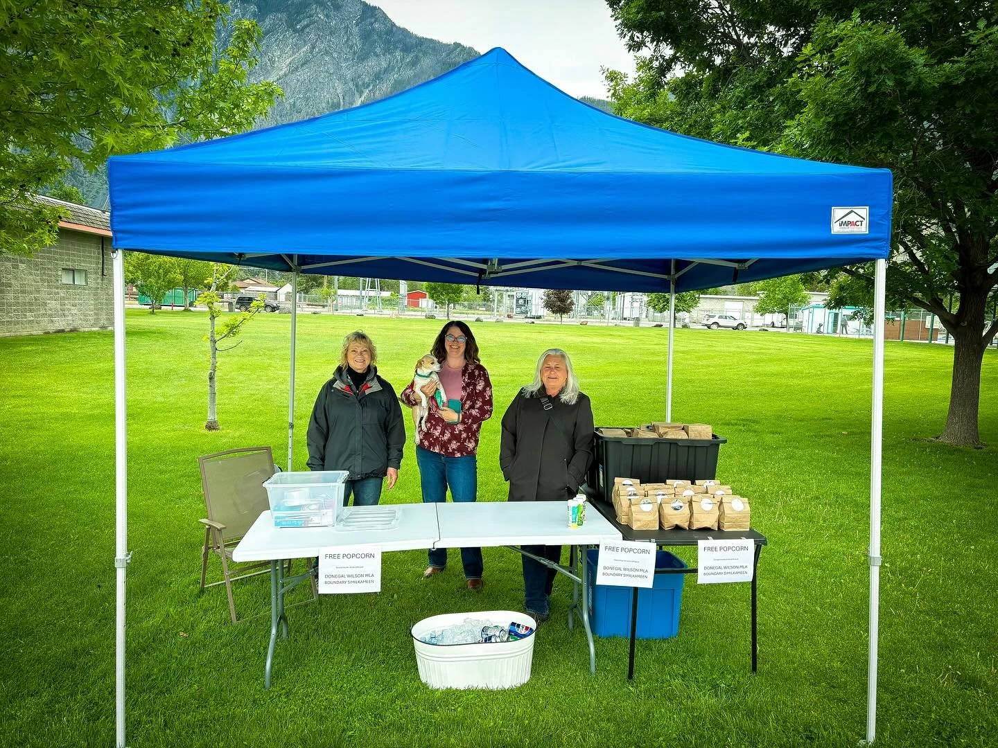 Donegal participating in the Keremeos Parade festivities with tables offering free popcorn, surrounded by trees and green grass.