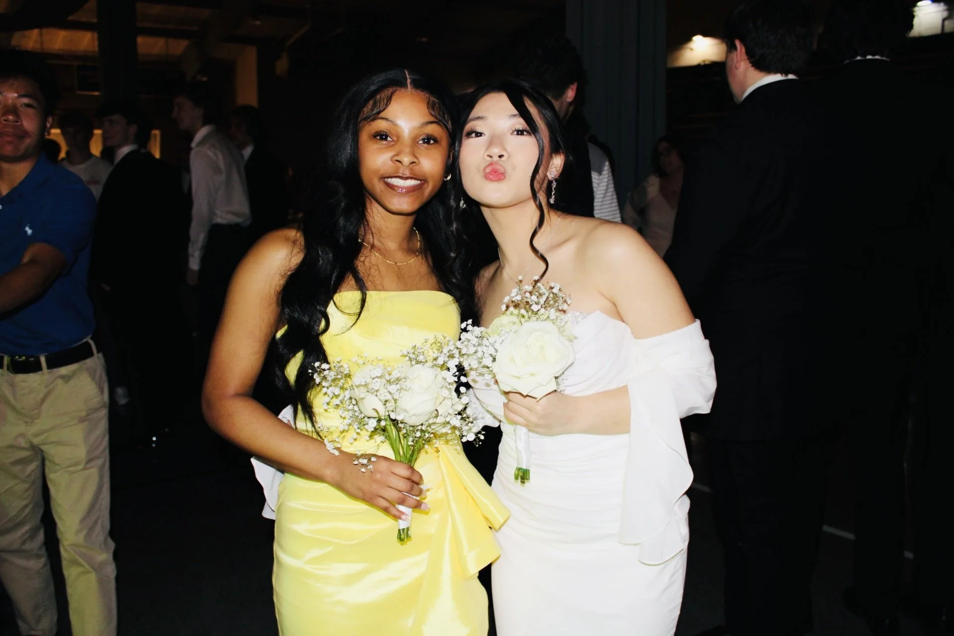 Two women at a formal event, one in a bright yellow dress and the other in a white dress, both holding bouquets of flowers, with other people in the background.