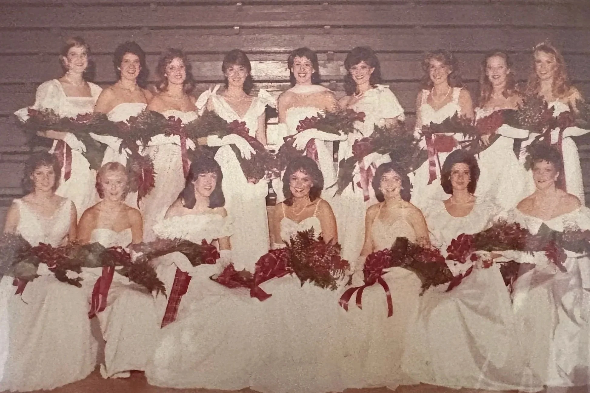 Vintage photo of twenty women in white dresses with red ribbons, holding bouquets, posed in two rows against a wooden wall.