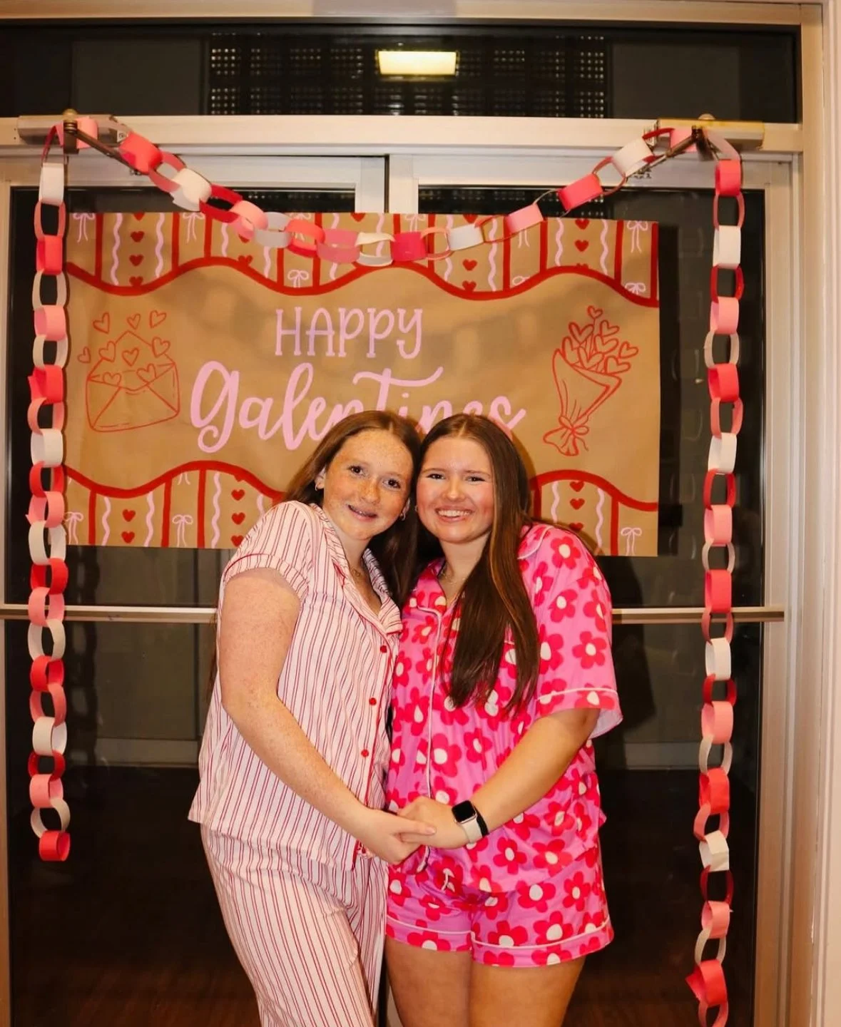 Two young women in pajamas, holding hands and smiling in front of a Valentine's Day themed backdrop with the words 'Happy Galentine's' and decorations of hearts, envelopes, and a bouquet.