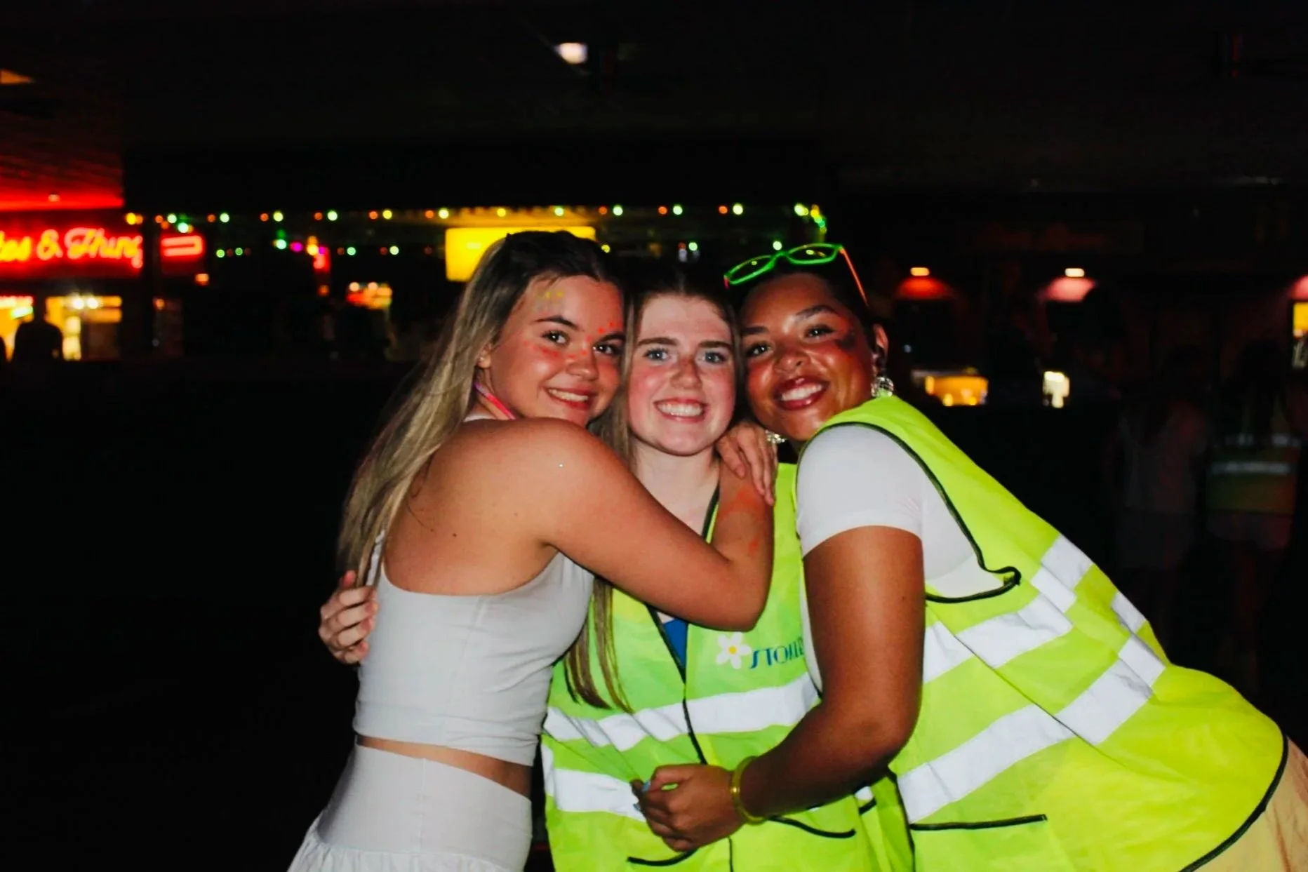 Three women smiling and hugging each other in a dimly lit indoor setting with colorful string lights in the background. Two women are wearing reflective safety vests.