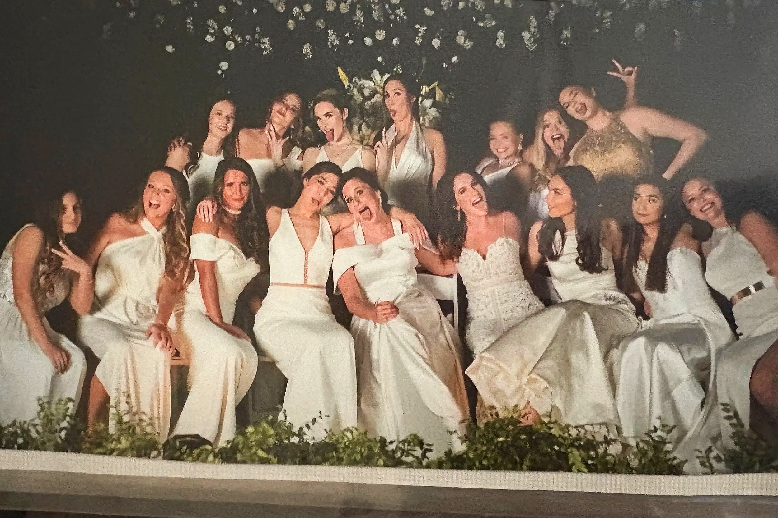 A group of women dressed in white wedding gowns and formal wear, smiling and posing together at a wedding or celebration, with a dark background and floral decorations.