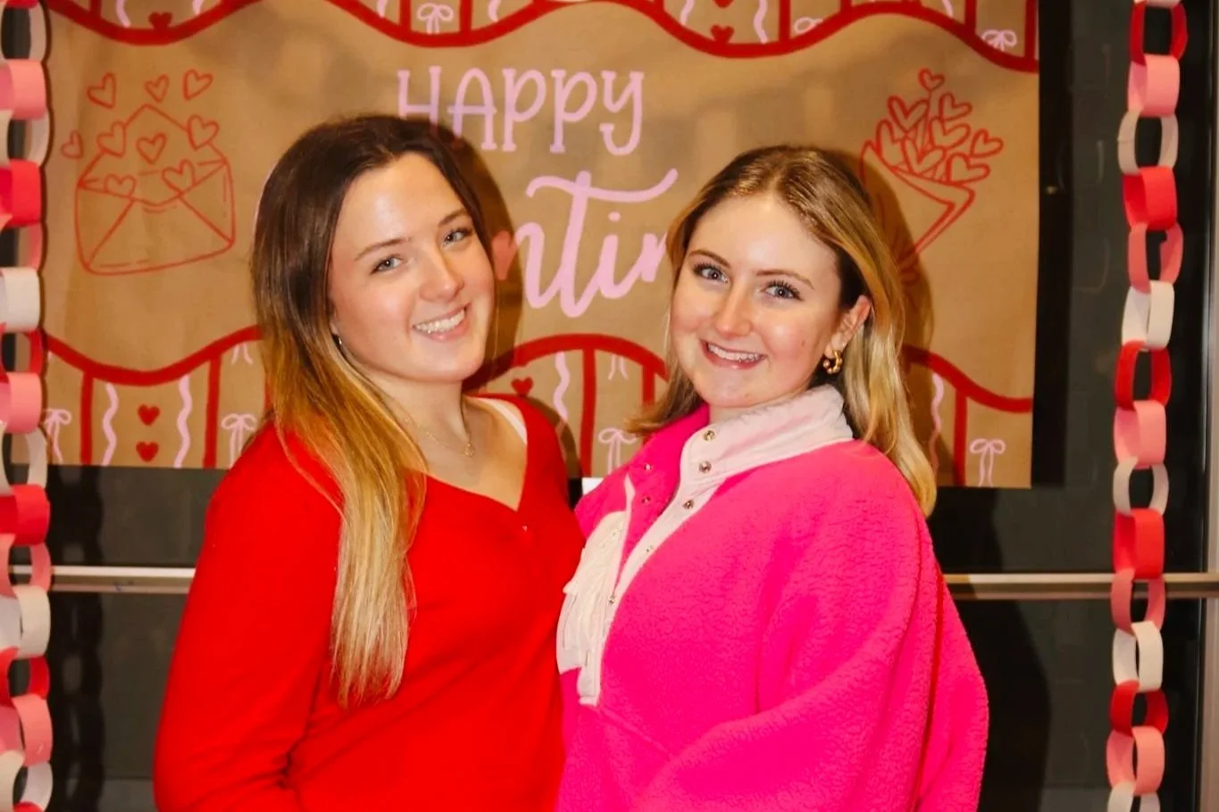 Two women smiling at a Valentine's Day celebration, standing in front of a decorated backdrop with hearts and the words "Happy Valentine". One woman is wearing a red top, and the other is dressed in a pink fleece jacket.
