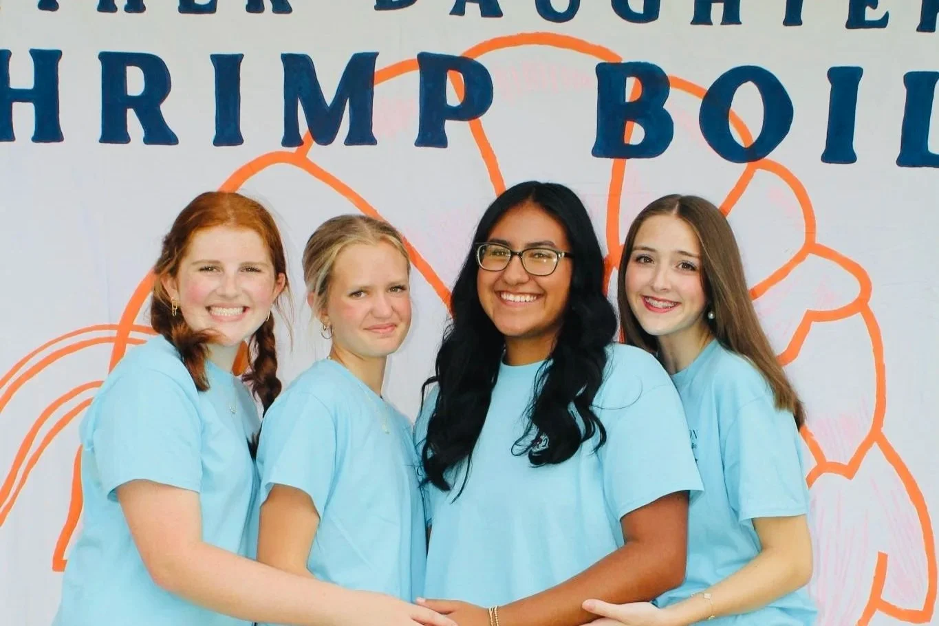 Four young women wearing matching light blue T-shirts standing together and smiling, with a graphic background featuring shrimp and the words "Shrimp Boil".