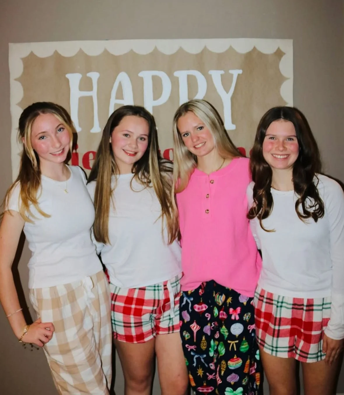 Four young women standing together at a birthday celebration, smiling in front of a beige backdrop with white and red lettering that says 'HAPPY'.