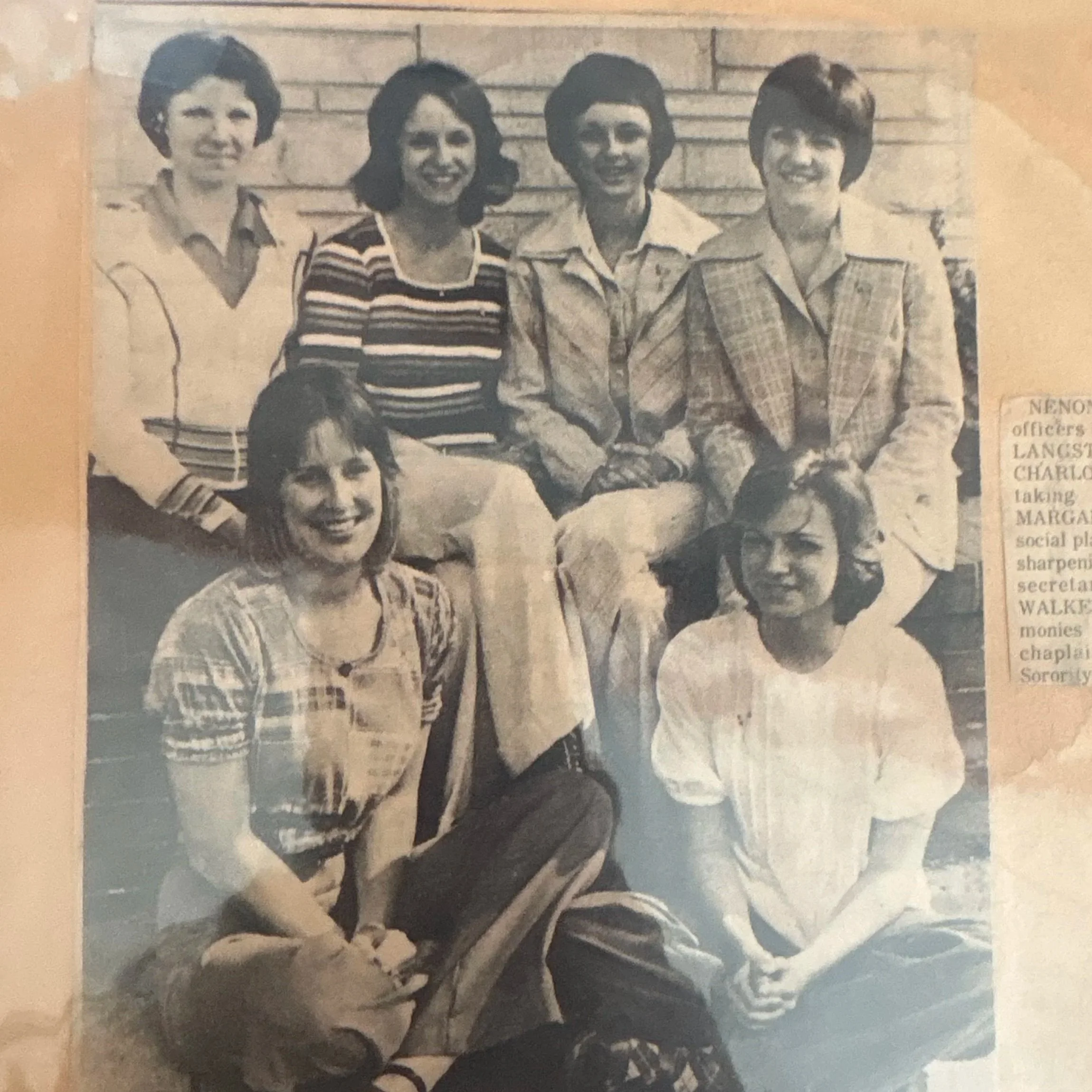Group of seven women posing outdoors in front of a brick wall, some sitting and some standing, in vintage clothing from the late 20th century.