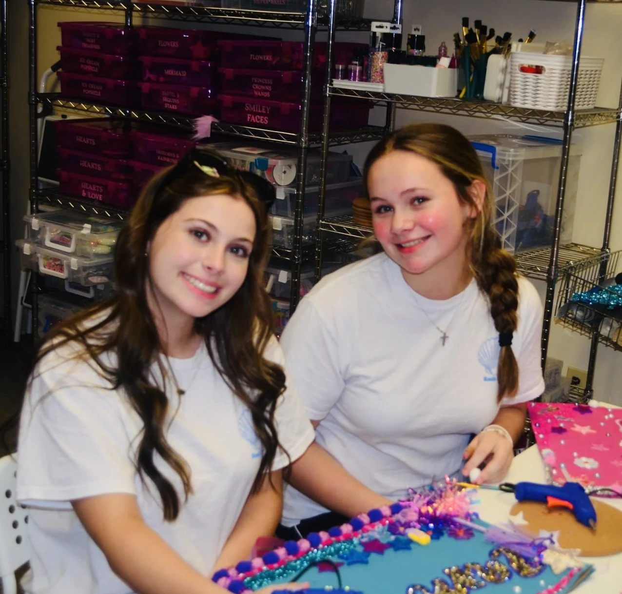 Two young girls sitting at a table working on a colorful craft project, surrounded by crafting supplies in a room with shelves filled with boxes and supplies.
