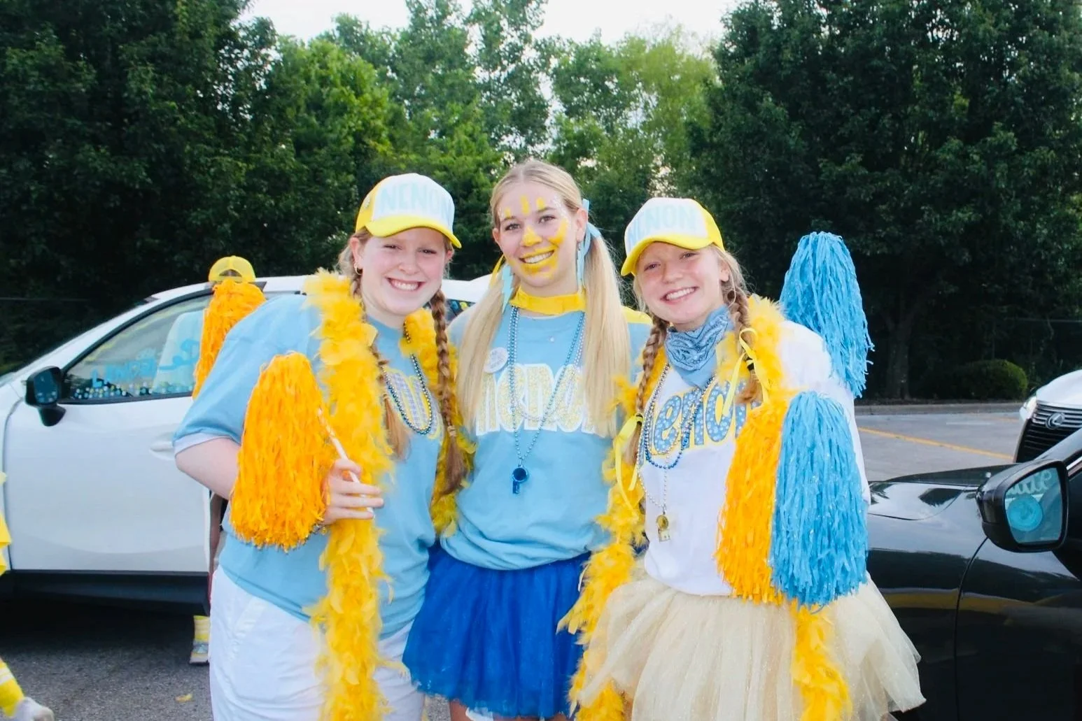 Three young girls dressed in blue and yellow outfits standing close together and smiling outside in a parking lot, with trees in the background.