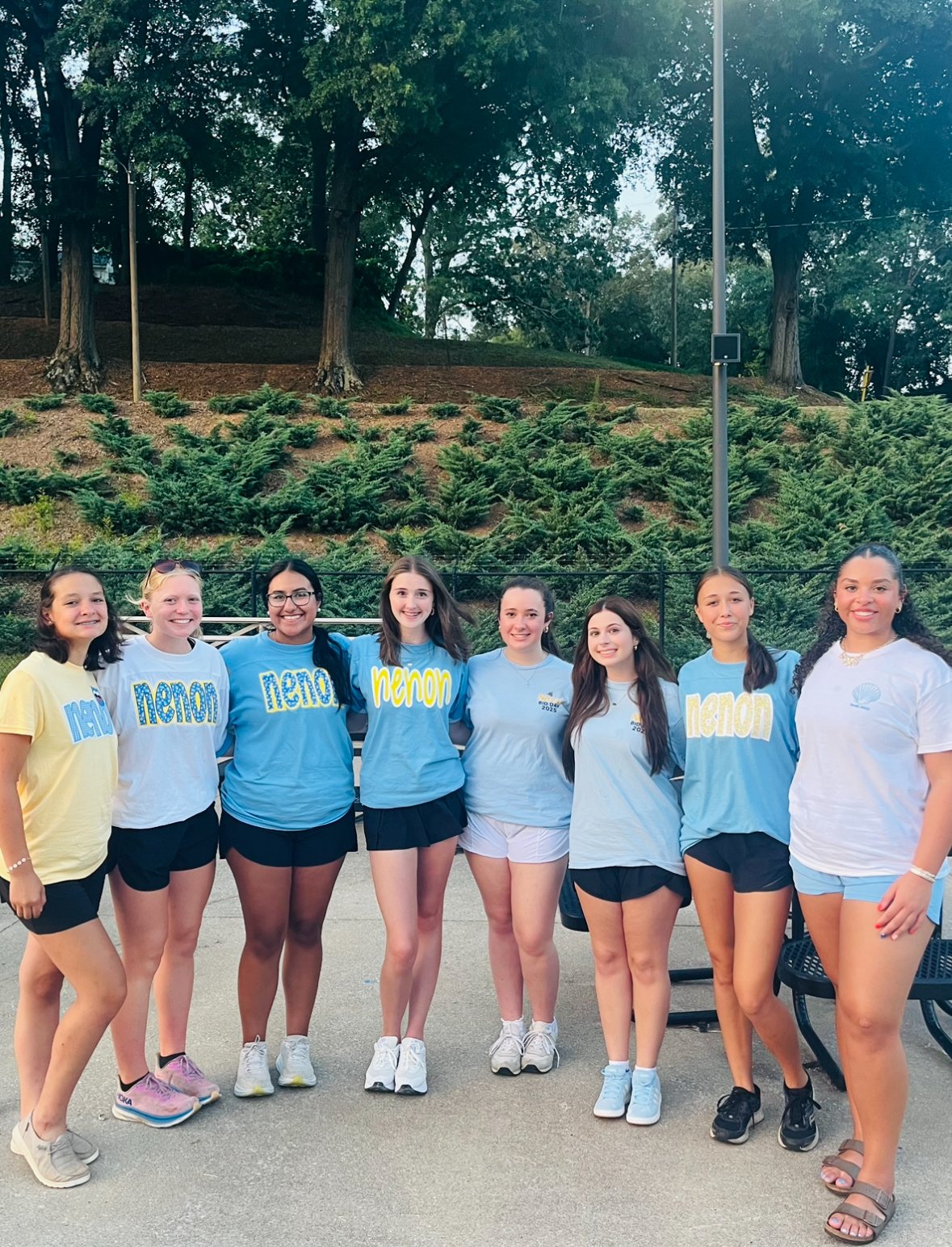 Group of eight young women standing together outdoors, wearing casual clothes and sportswear, smiling at the camera, with greenery and trees in the background.