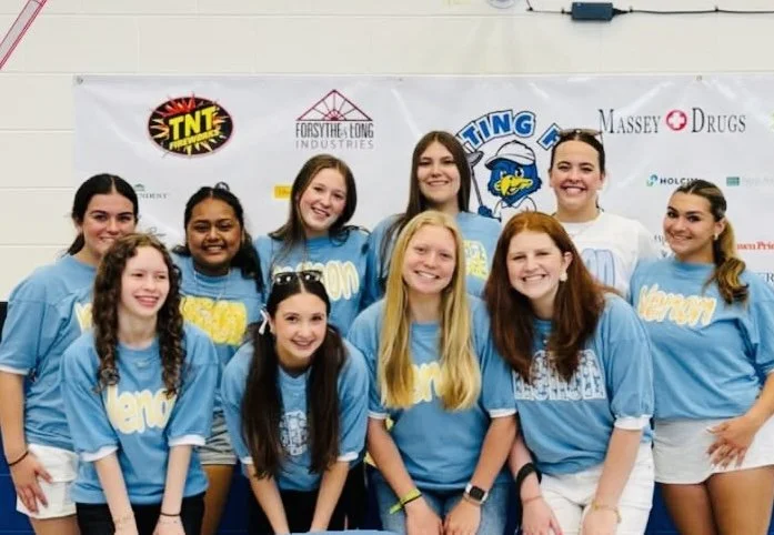Group of young girls in blue t-shirts posing for a photo indoors, with a banner featuring various sponsor logos in the background.