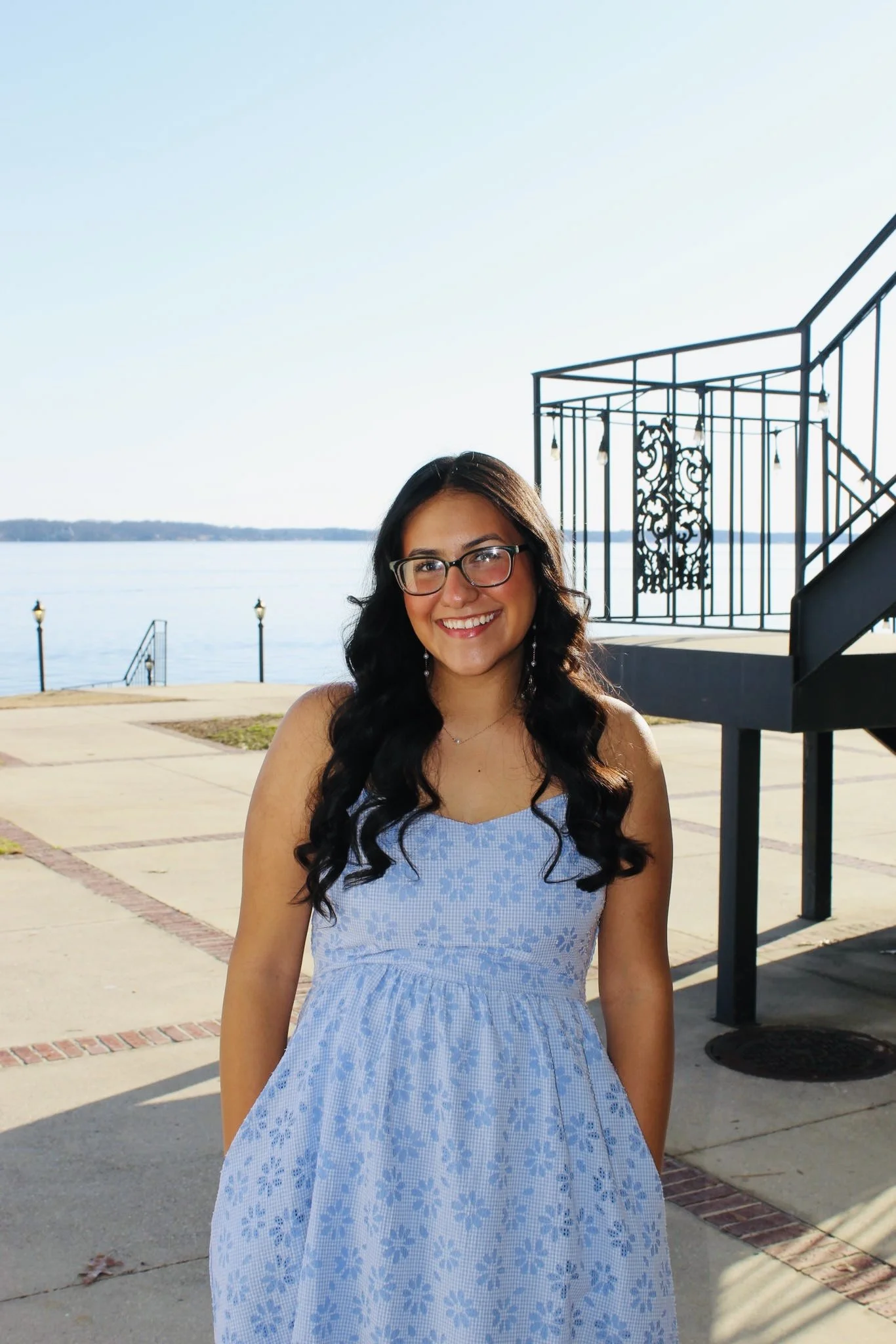 A young woman with long dark wavy hair and glasses is smiling outdoors near a body of water, wearing a light blue dress with a floral pattern.