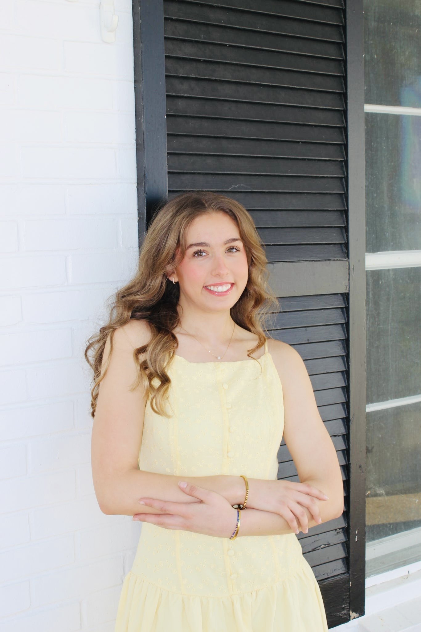 A young woman in a yellow dress smiling, standing outdoors near a black window shutter and a white brick wall.