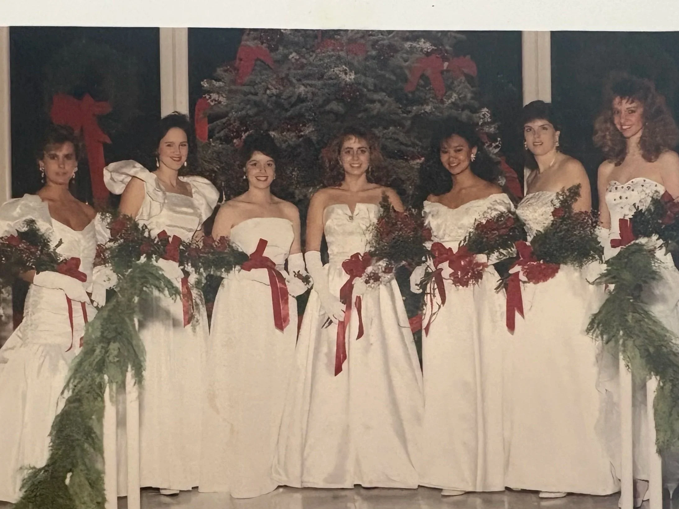 Seven women in white dresses holding bouquets of flowers with red ribbons, standing in front of a Christmas tree decorated with red bows.