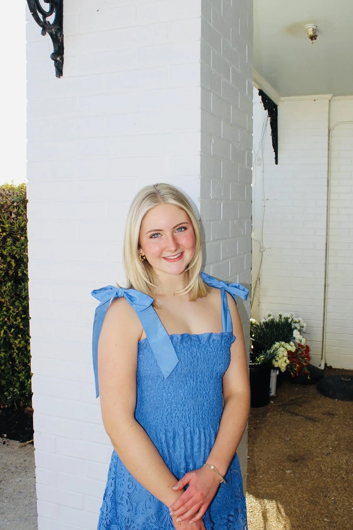 A young woman with blonde hair smiling and wearing a blue dress with bow shoulder straps standing outdoors against a white brick wall.
