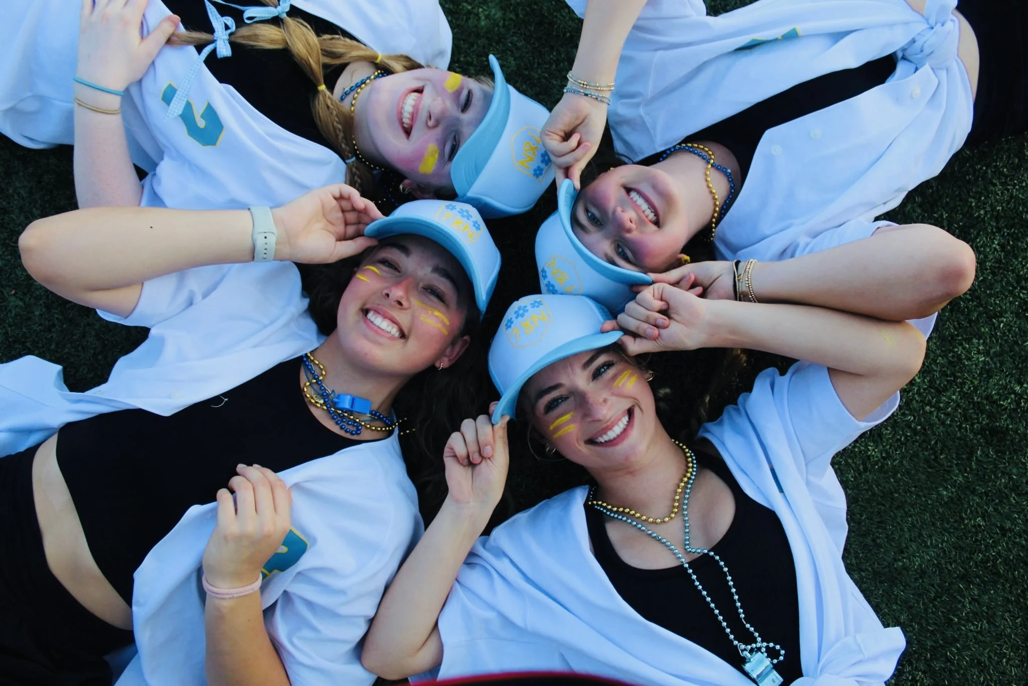 Group of five young women lying on the grass, wearing white and black clothing, blue caps, and decorated with yellow and blue face paint and accessories, smiling and looking up at the camera.