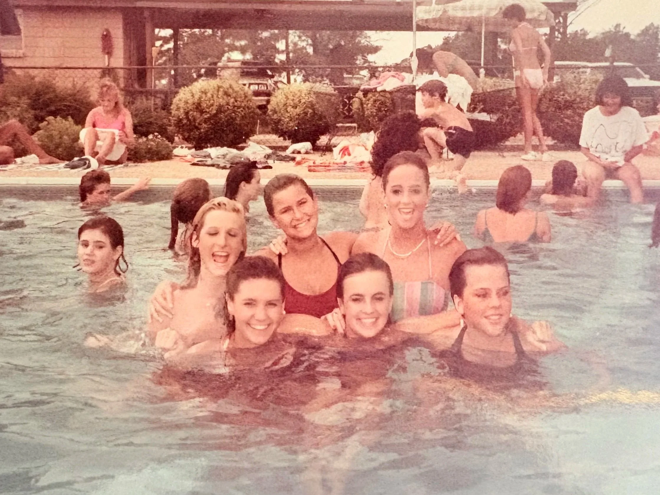 A group of seven women are in a swimming pool, smiling and posing for the photo with their arms around each other.