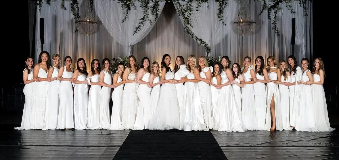 Group of women dressed in white gowns standing hand-in-hand on a stage with a draped white backdrop and chandeliers.