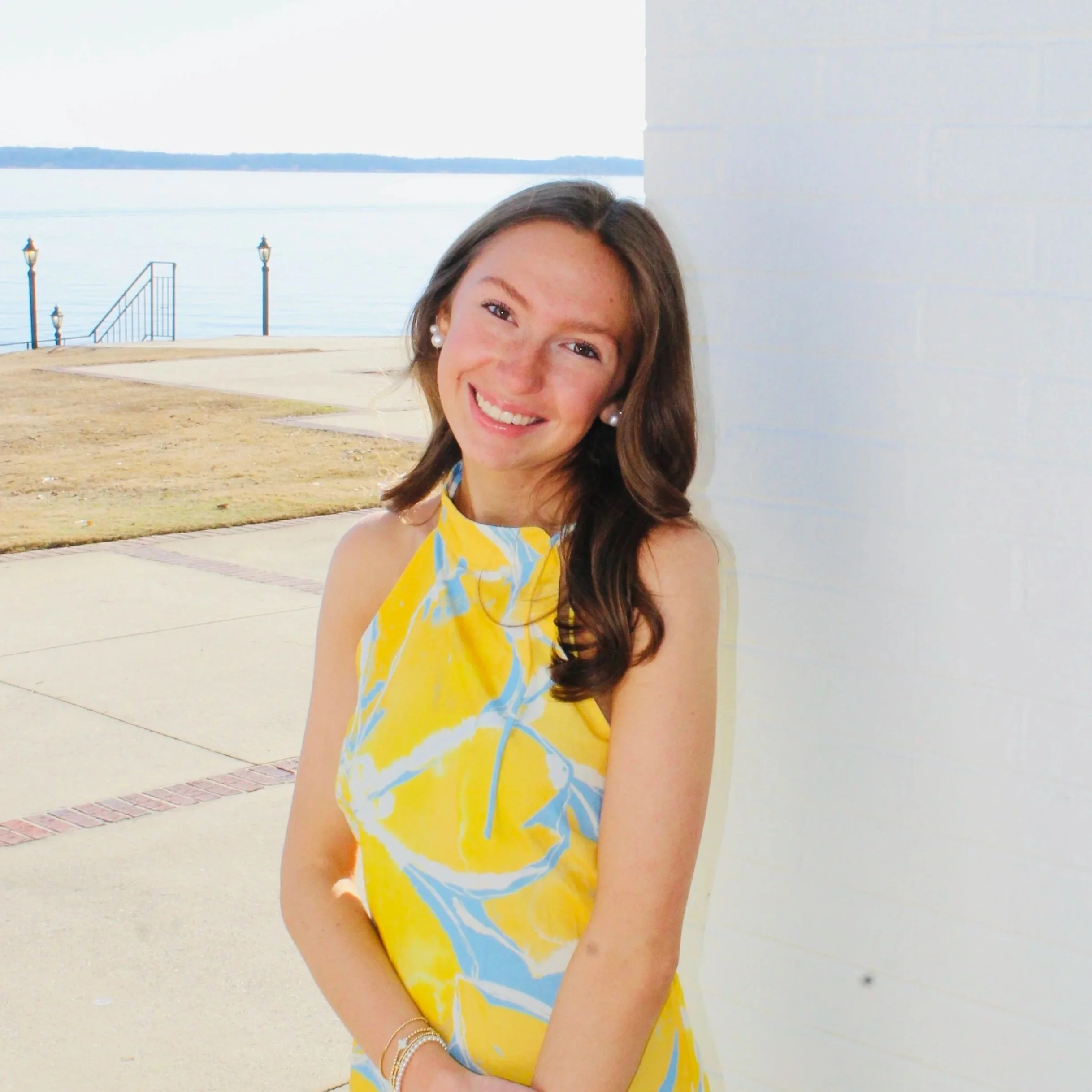Smiling young woman with long brown hair, wearing a yellow and blue patterned sleeveless dress, standing outdoors near a white wall with a body of water in the background.