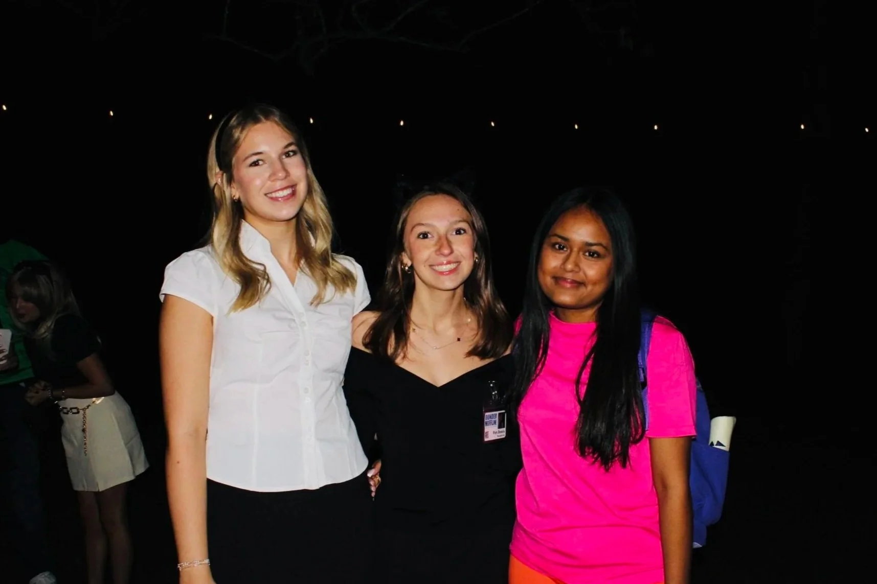 Three women posing together at night with dark background and small lights overhead. The woman on the left has blonde hair and is wearing a white short-sleeved top. The woman in the middle has brown hair and is wearing a black top. The woman on the right has long black hair and is wearing a pink t-shirt with orange shorts and a blue backpack.