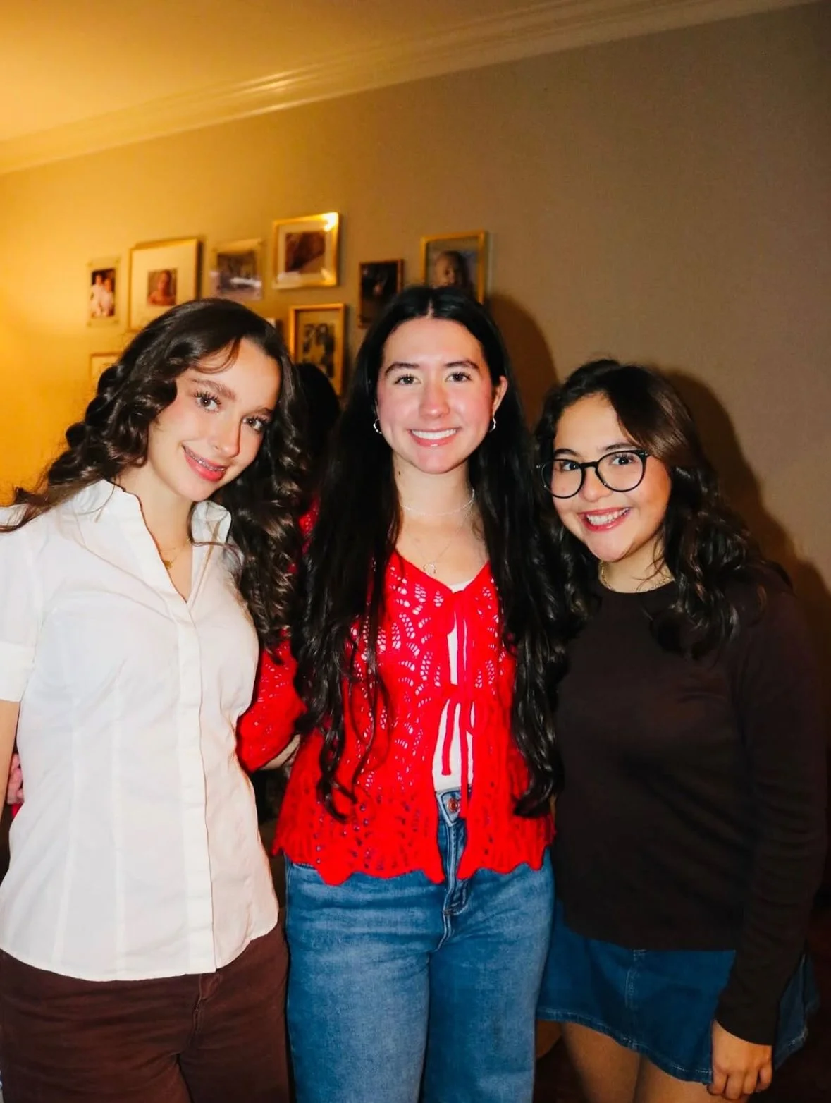 Three young women smiling and standing close together indoors, with framed photos on the wall behind them.