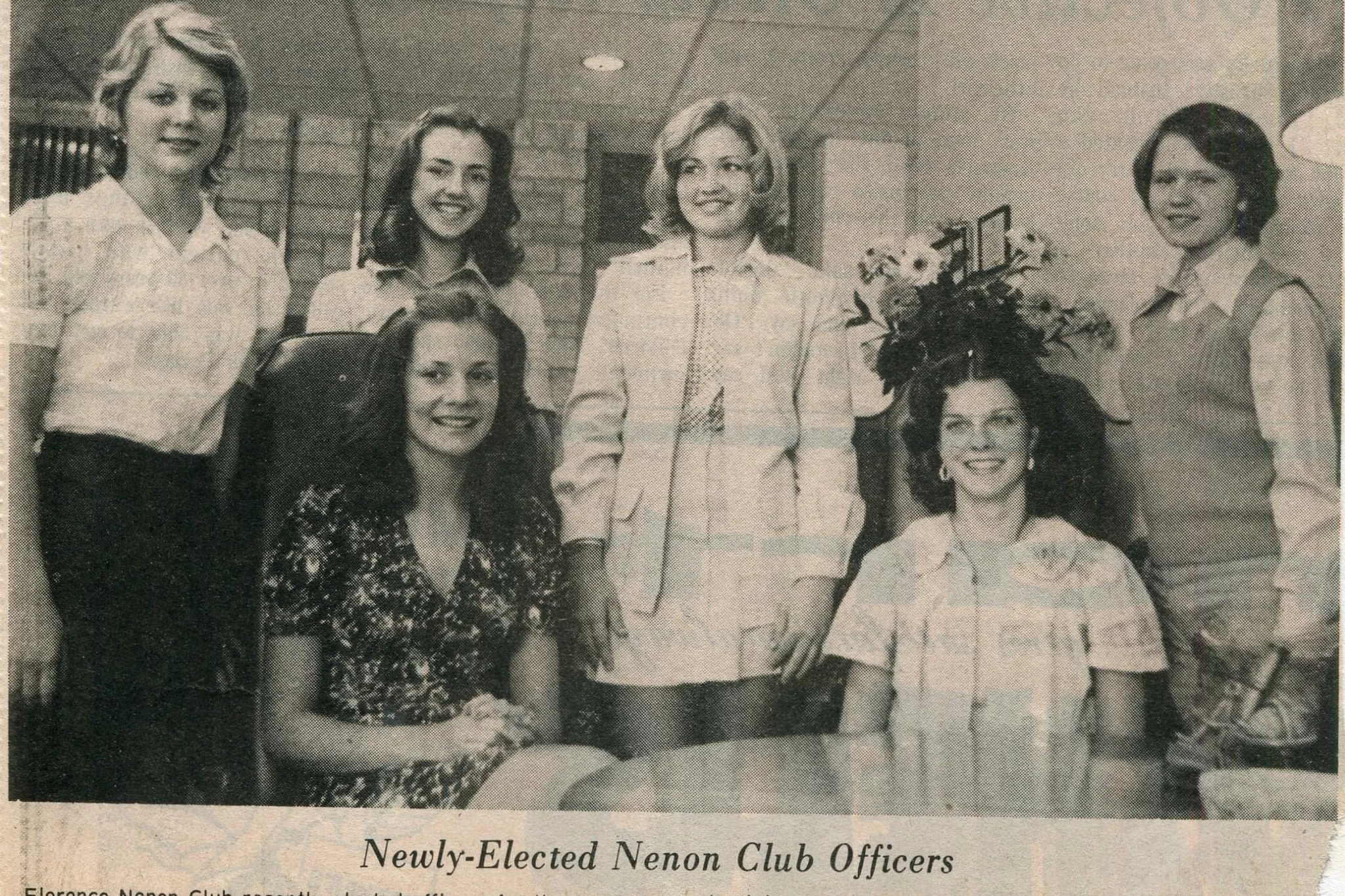 Group of seven women, some standing and some seated, posing in an indoor setting. The woman in the center has a large floral arrangement on her head, and others are smiling. The scene appears to be a formal or celebratory event.