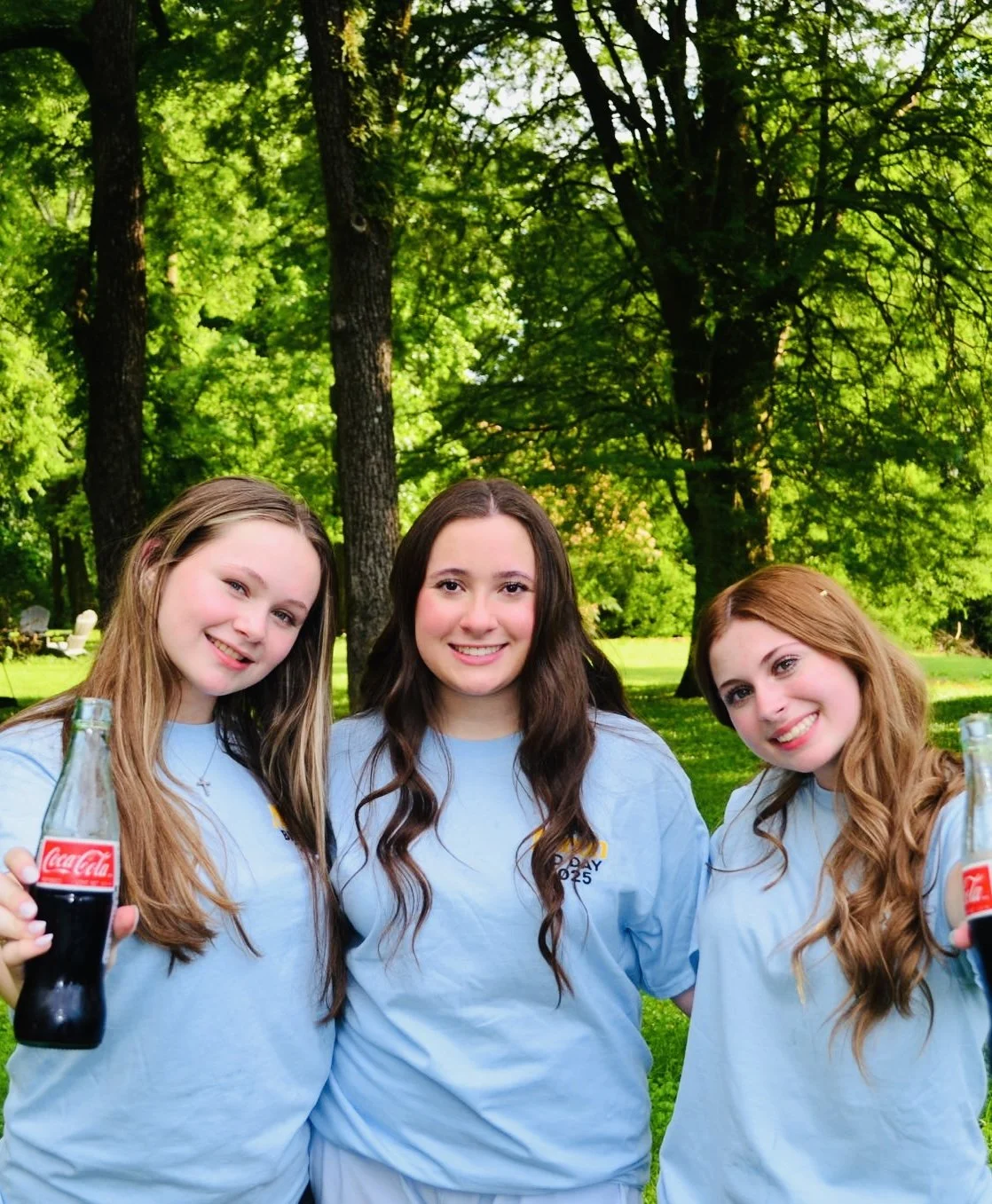 Three young women standing outdoors in a park with tall trees and green foliage, smiling and holding bottles of Coca-Cola, wearing matching light blue T-shirts.